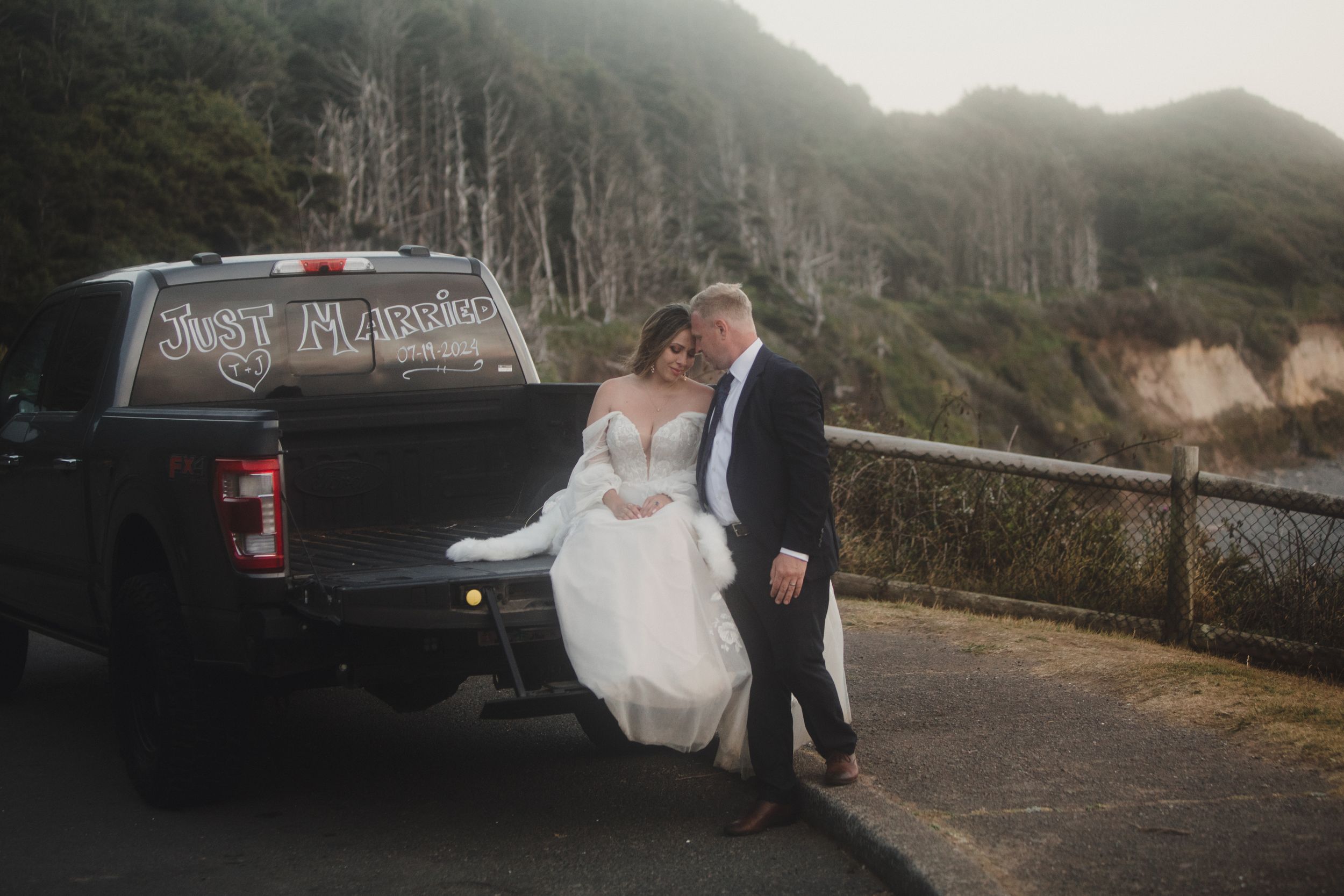 Wedding party poses near vintage truck on foggy coastal road with dramatic cliffside views.