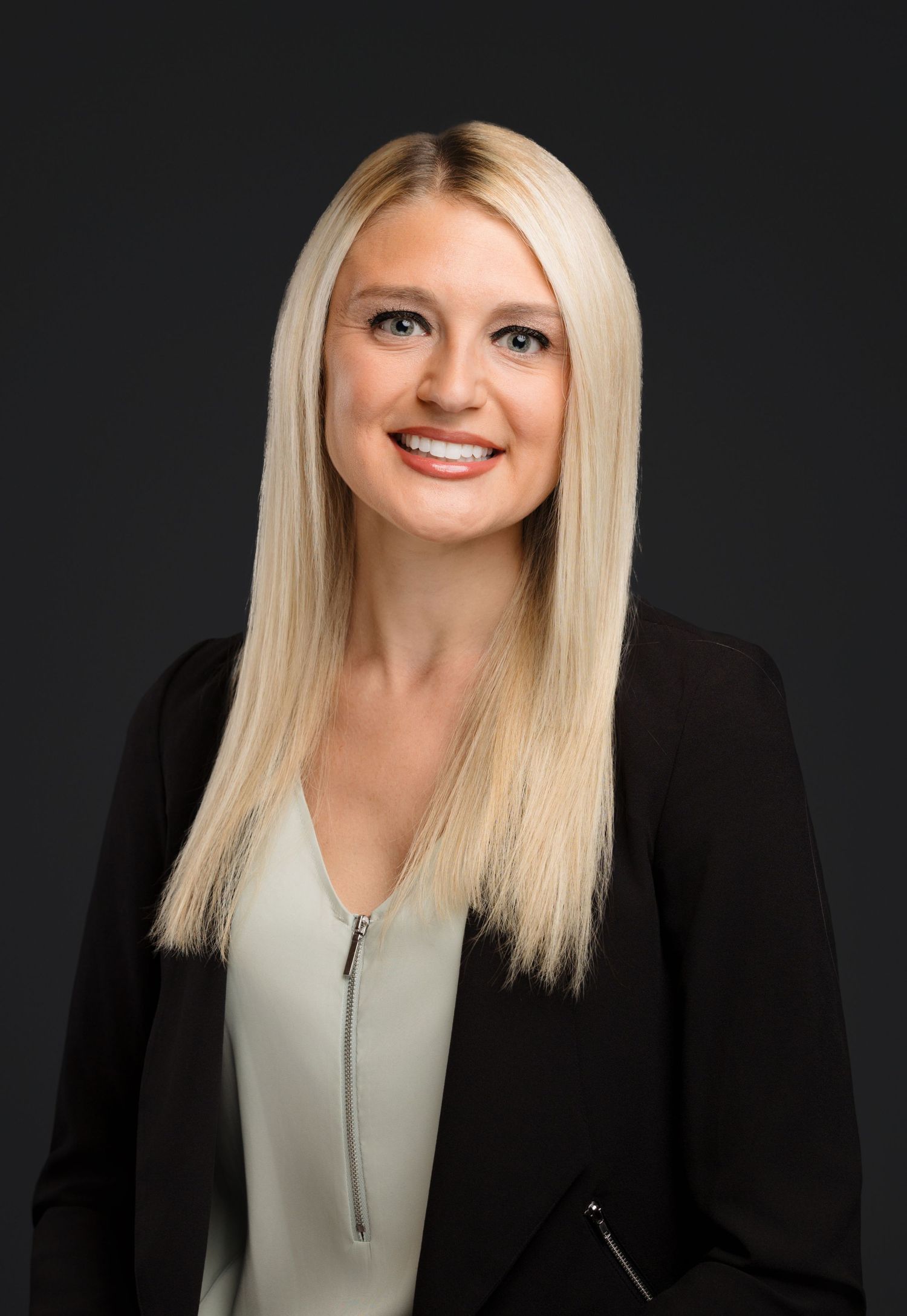 Professional headshot portrait with blonde hair wearing black blazer and light colored top against dark background.