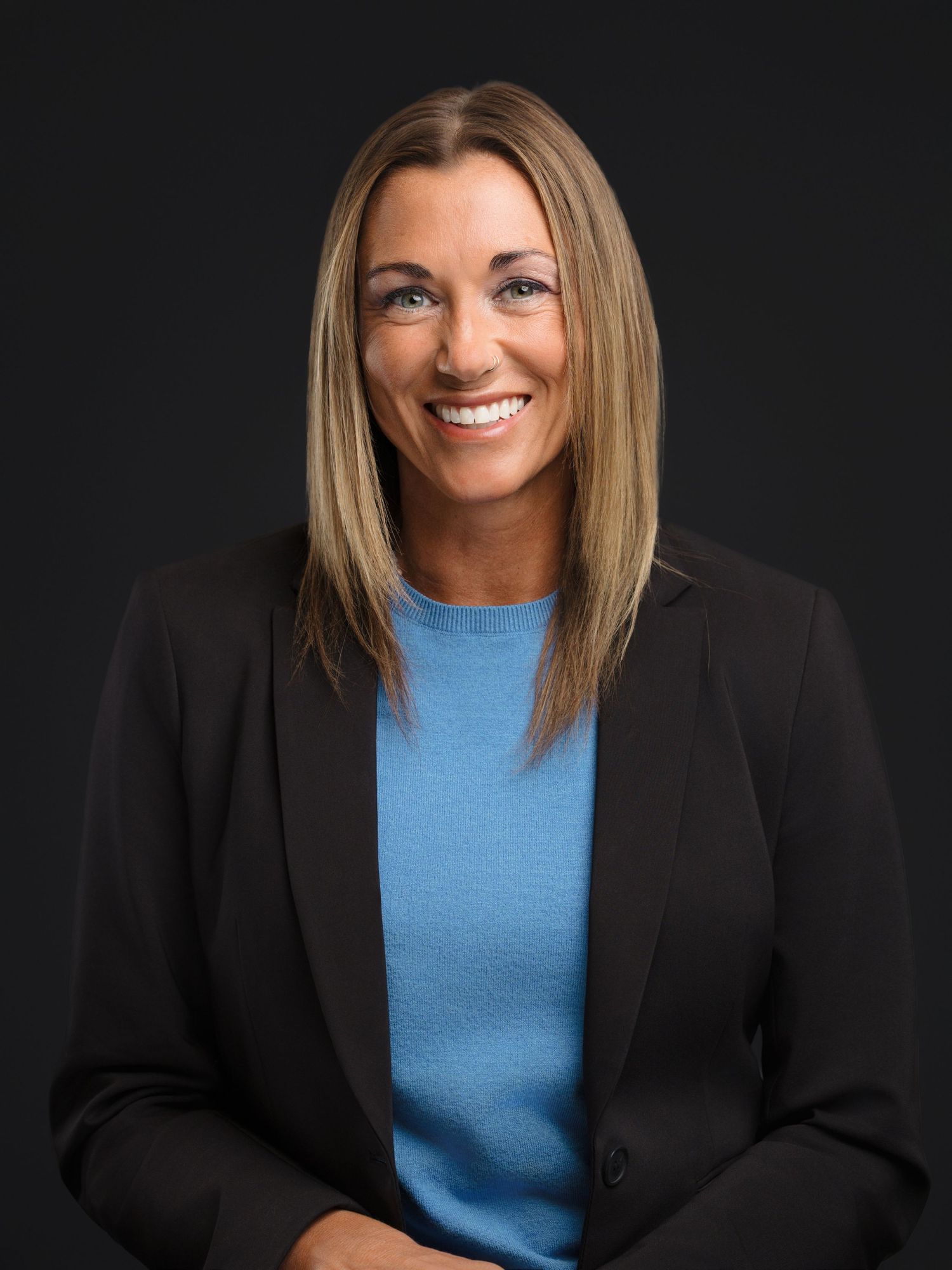 Professional headshot portrait in black blazer and blue top against dark background with warm expression.