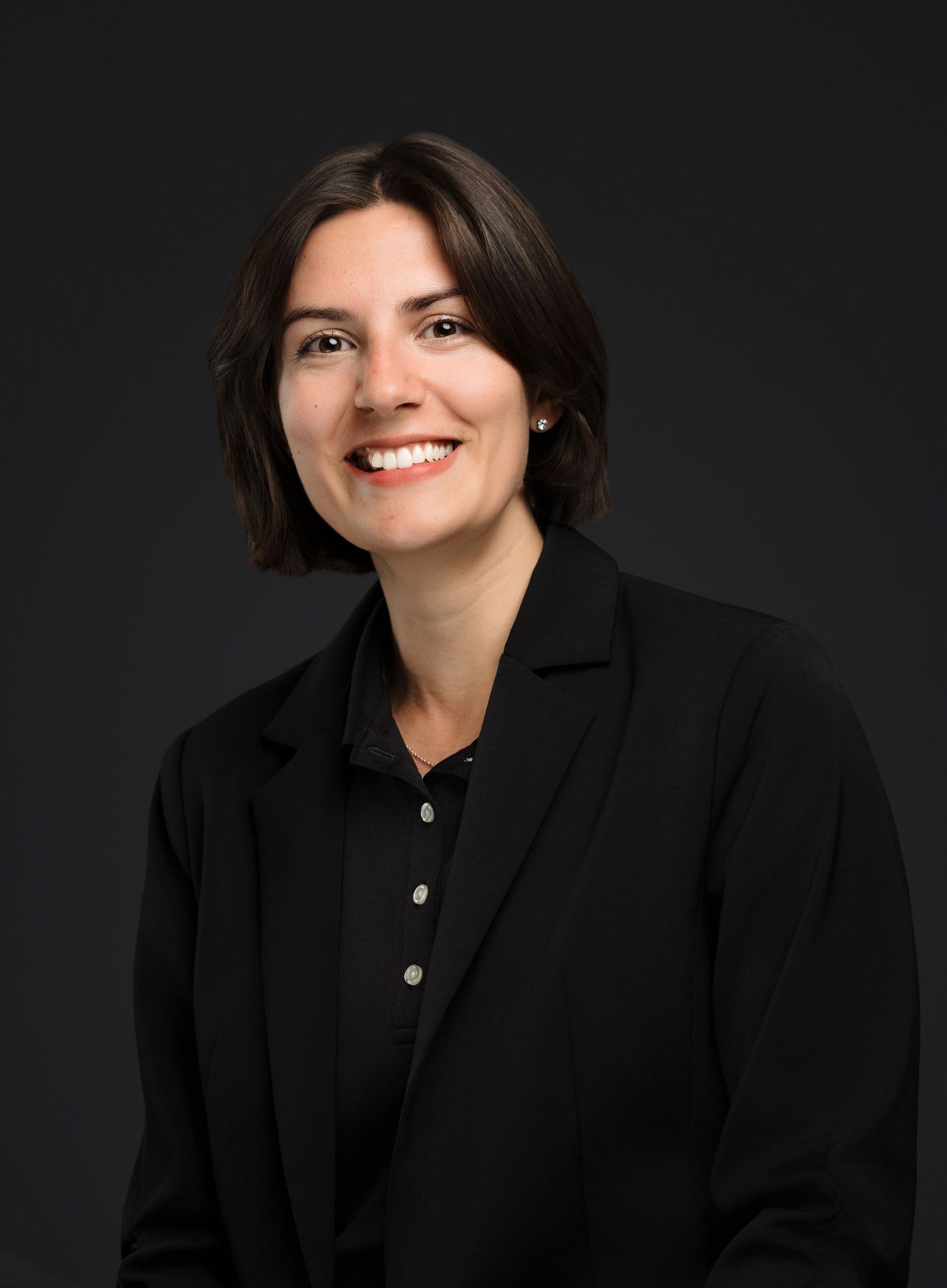 Professional headshot portrait of someone wearing a black blazer and polo shirt against a dark background.