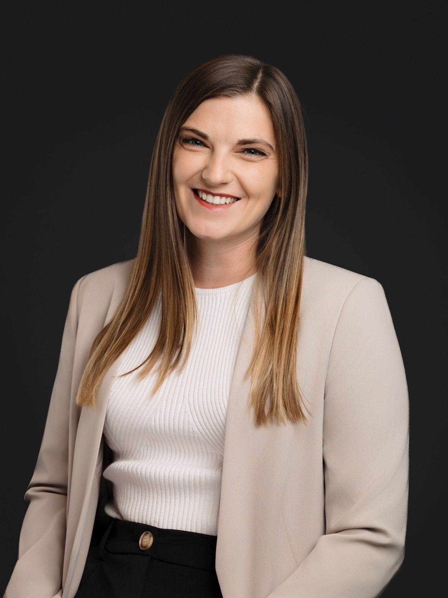 Professional studio portrait of someone wearing a white ribbed top and beige blazer against a dark background.