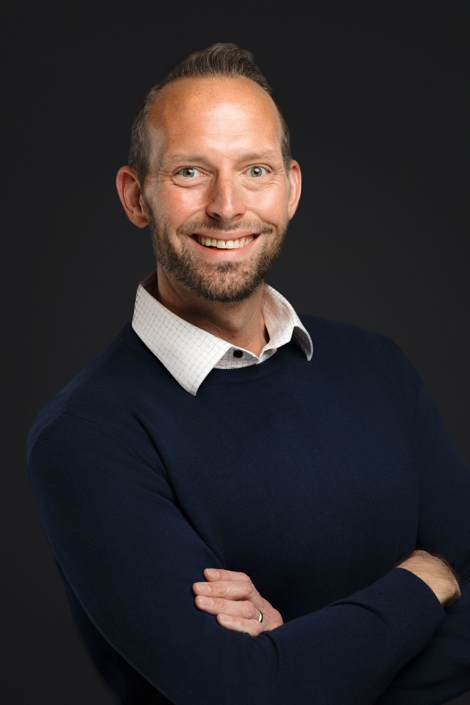 Professional studio portrait against dark background showing warm smile and navy sweater over collared shirt.