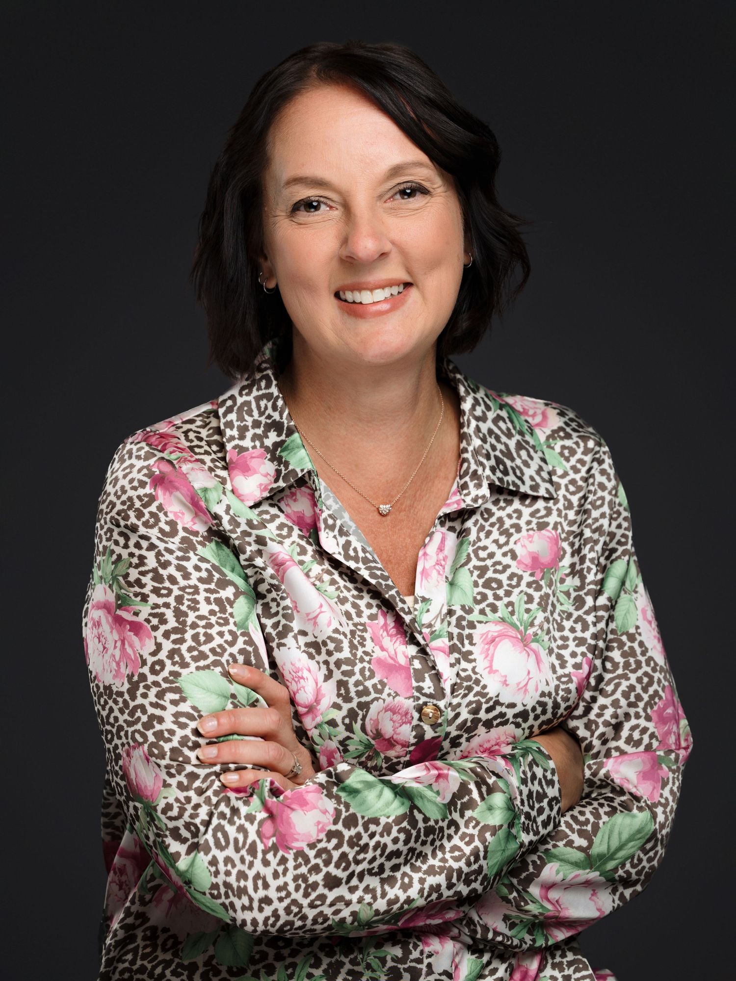 Professional headshot of someone wearing a floral and leopard print blouse with arms crossed against dark background.