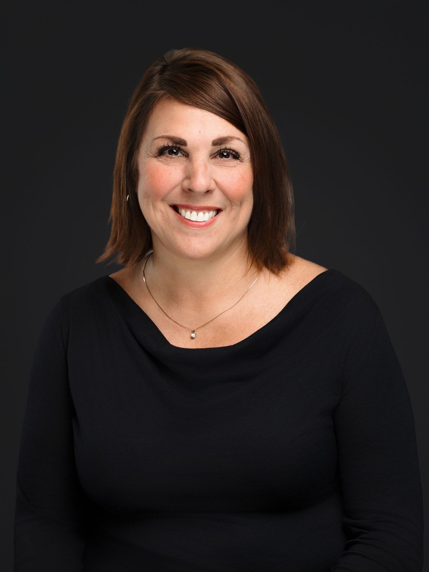 Professional headshot portrait of someone wearing a black top against a dark background, showing a warm smile.