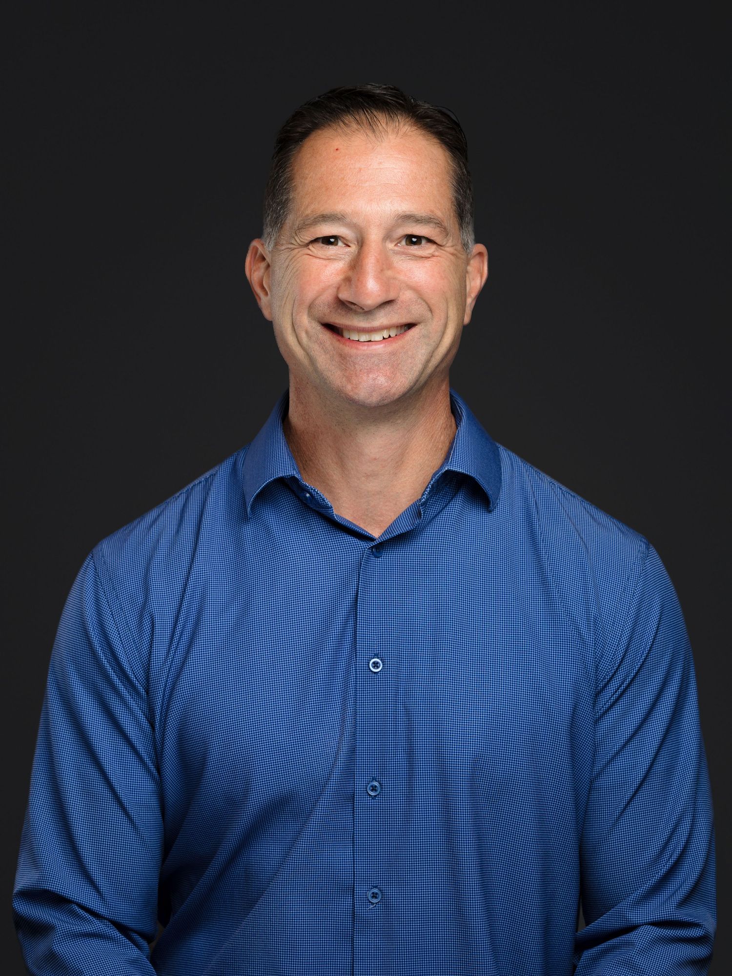 Professional headshot portrait of person in blue button-down shirt against dark background with friendly smile.