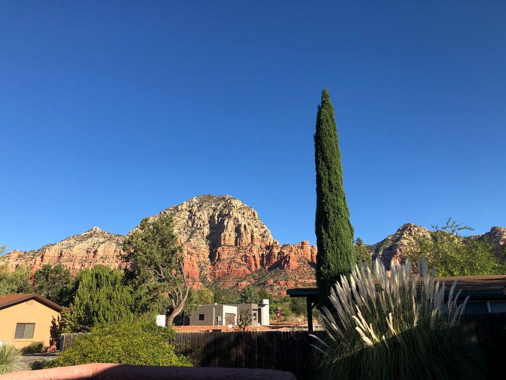 A panoramic view of red rock formations and desert landscape with cypress trees against a bright blue Arizona sky.