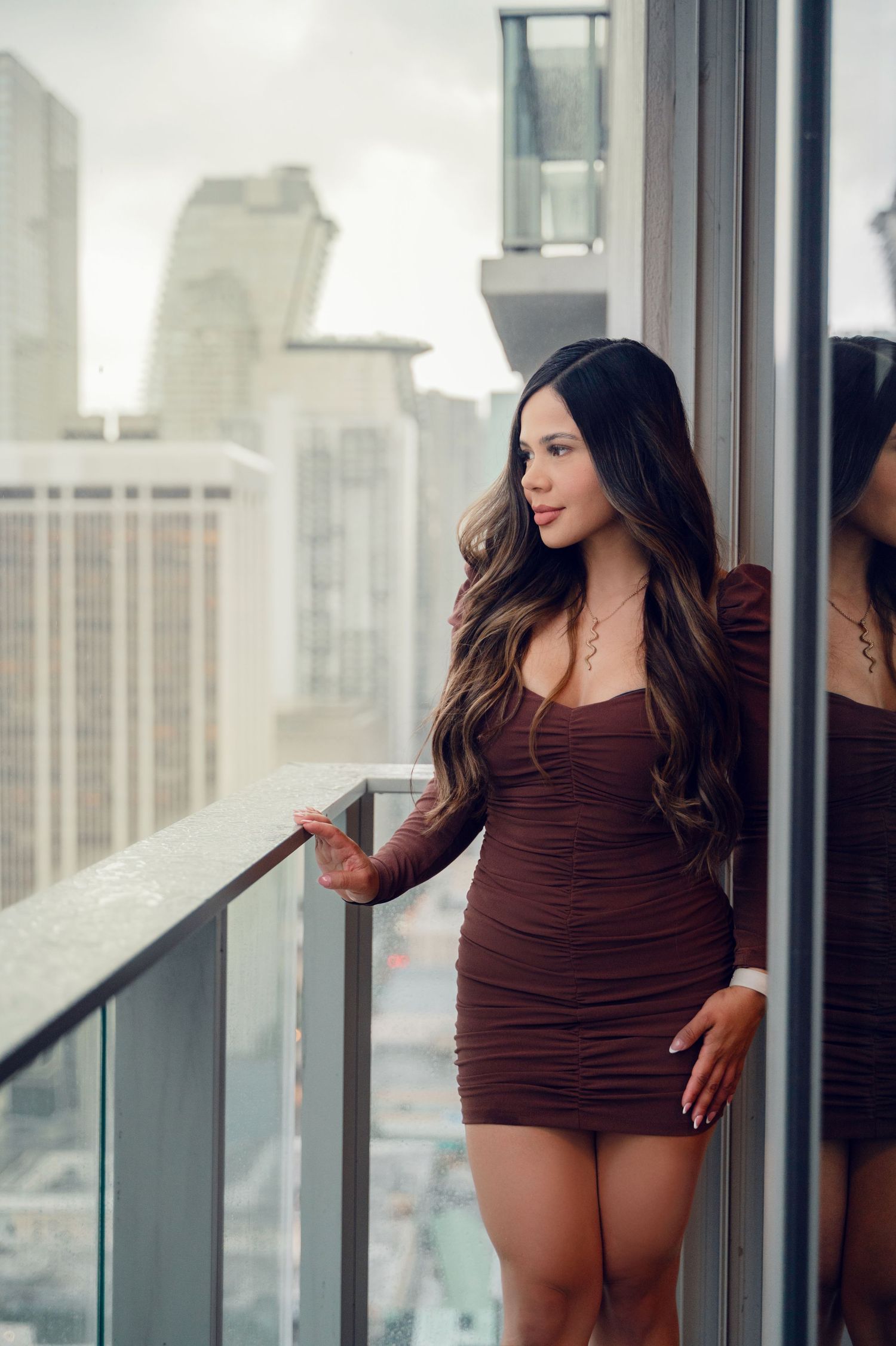 Woman in brown dress poses by window with city skyline view in background.