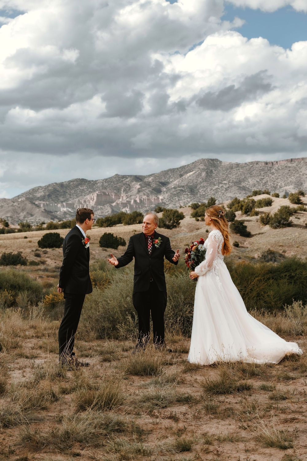 Bride and groom meeting at outdoor desert wedding ceremony with officiant, mountains and dramatic clouds in background.