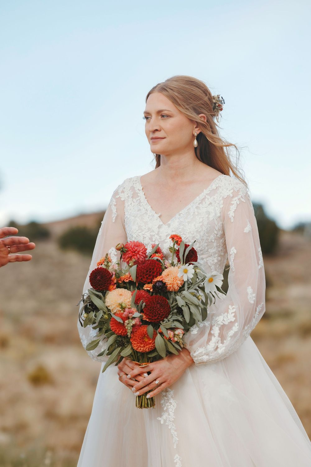 Elegant bride in lace long-sleeve wedding gown holding vibrant red and orange dahlia bouquet in outdoor desert setting.