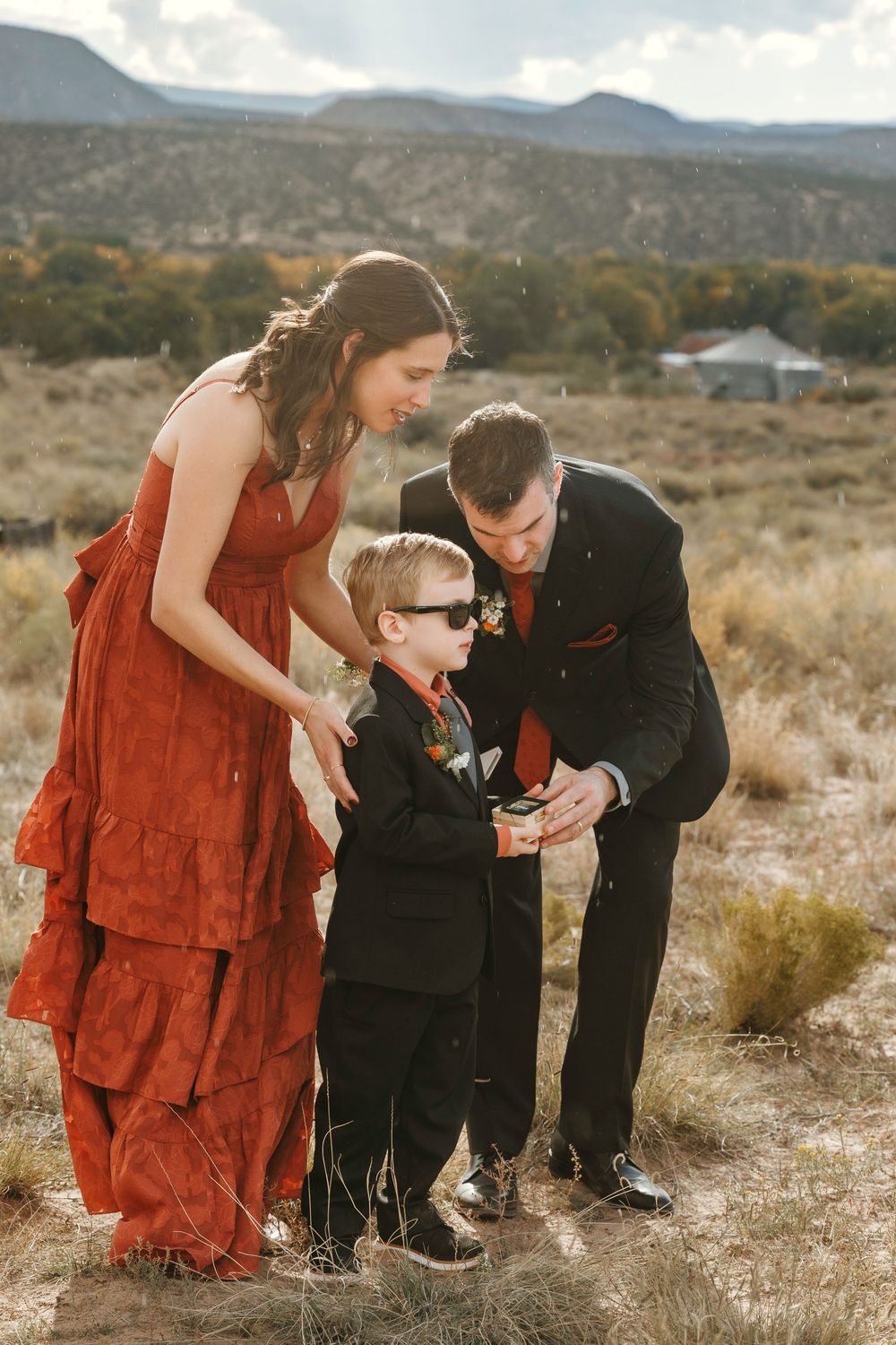 Woman in rust orange dress and man in black suit attending to young ring bearer boy in sunglasses at outdoor wedding.