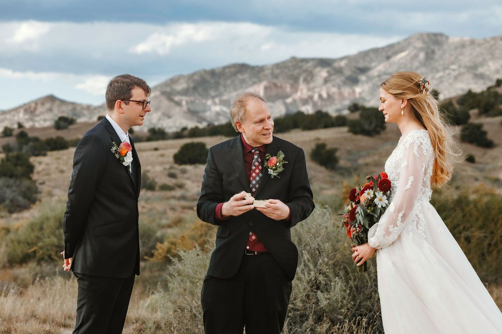 Wedding officiant reads vows as groom in black suit and bride in lace gown face each other in desert landscape.