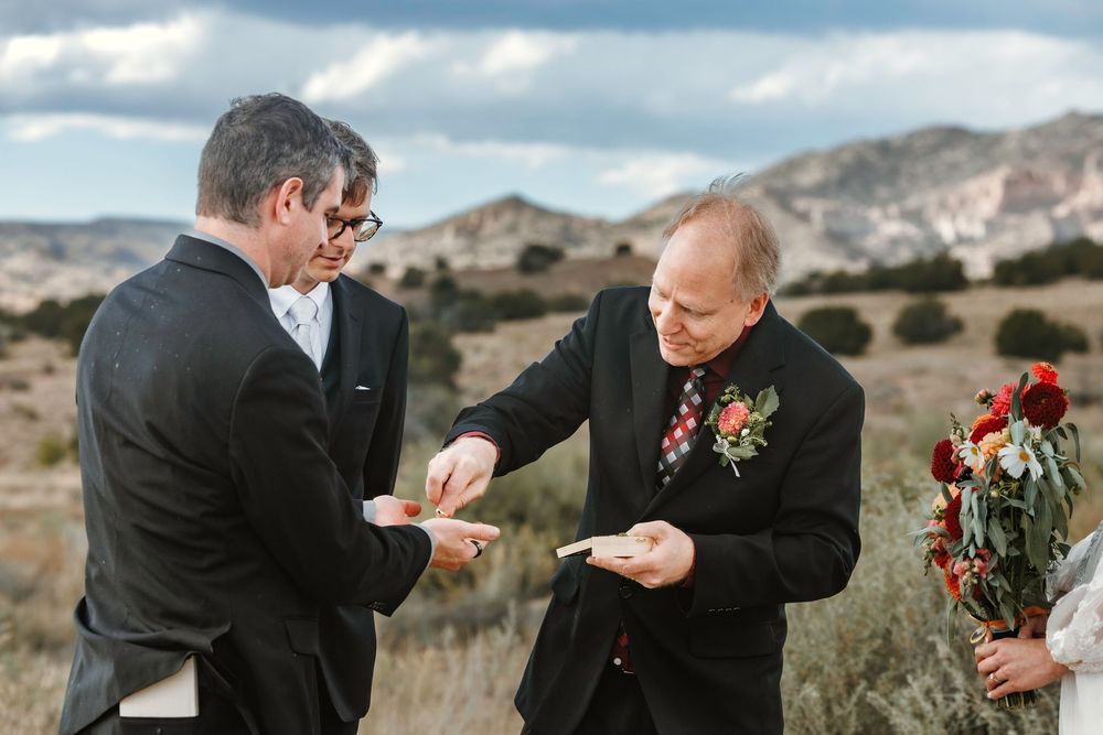 Groom and officiant exchange wedding rings during outdoor ceremony with arid desert hills in the background.