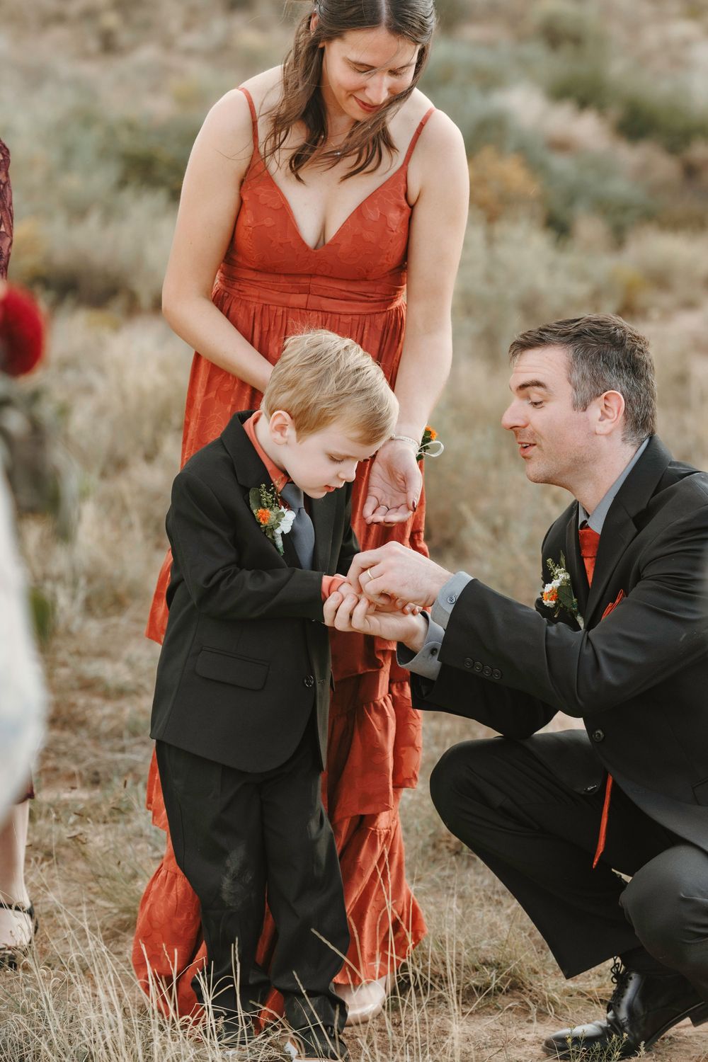 Smiling groomsman kneels to interact with young ring bearer boy while bridesmaid in rust dress looks on warmly.