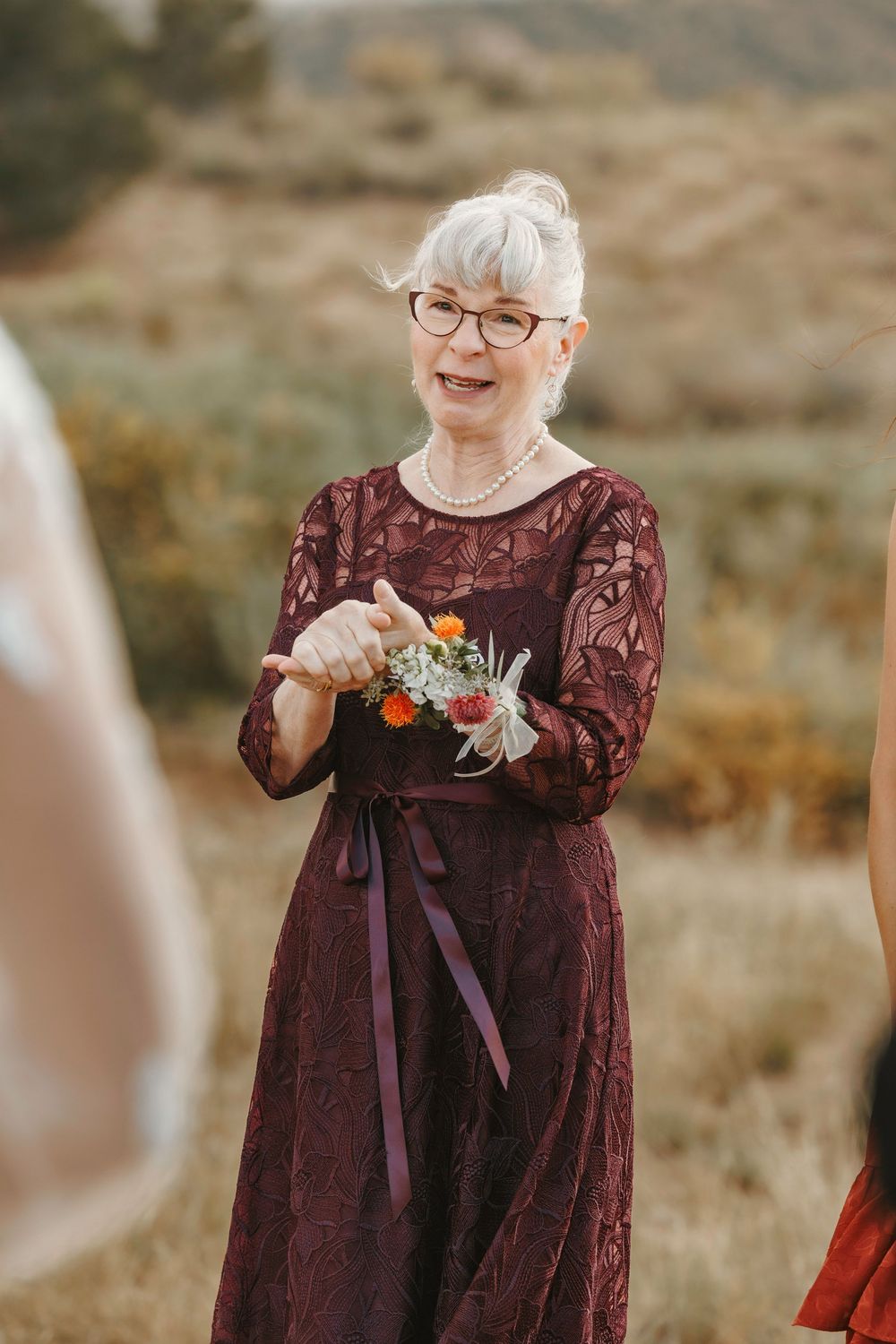 Joyful older woman in burgundy lace dress holding small floral bouquet smiling warmly at outdoor wedding ceremony.