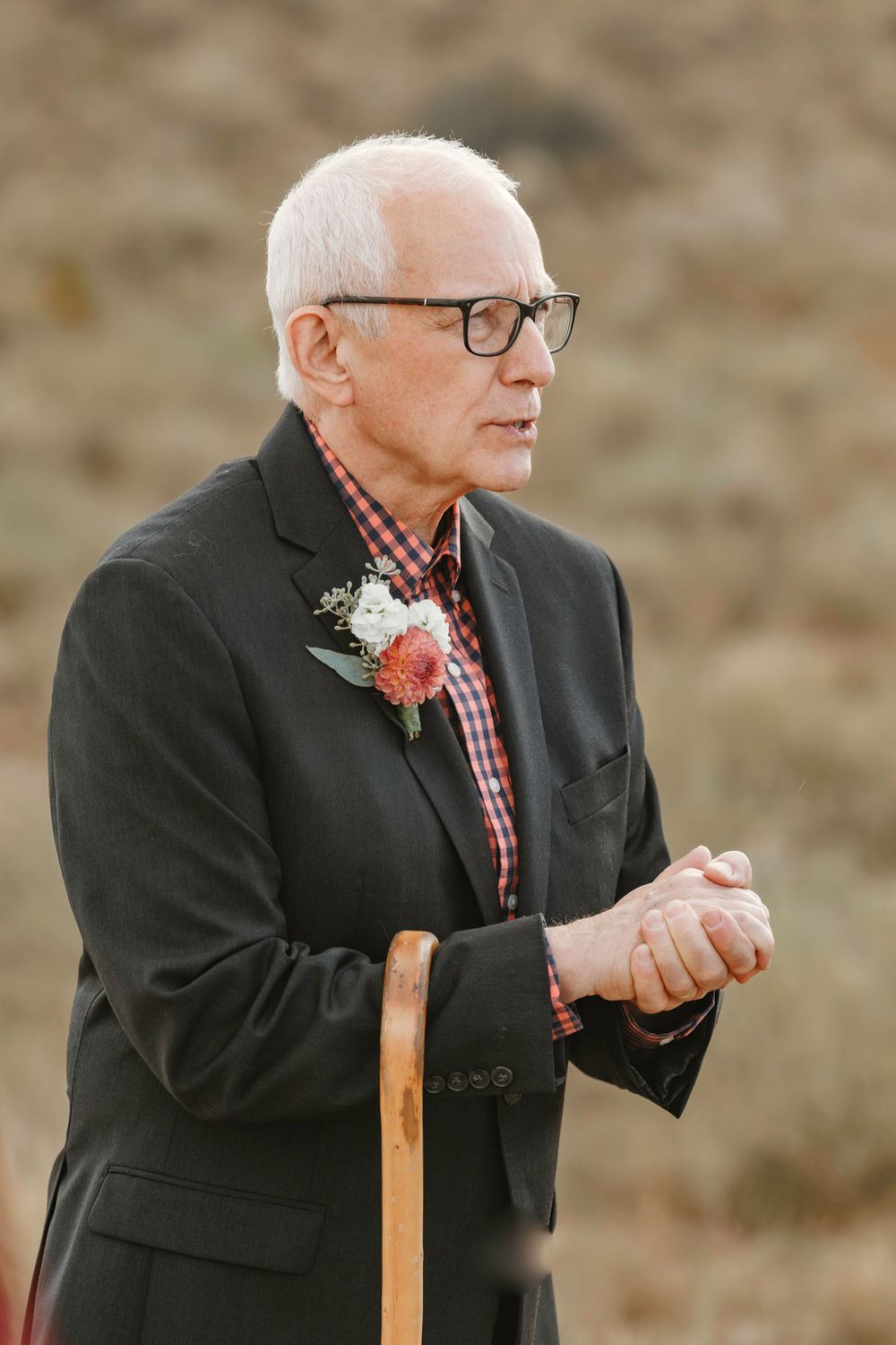 Elderly man in black suit with boutonniere leaning on wooden cane watching outdoor wedding ceremony attentively.