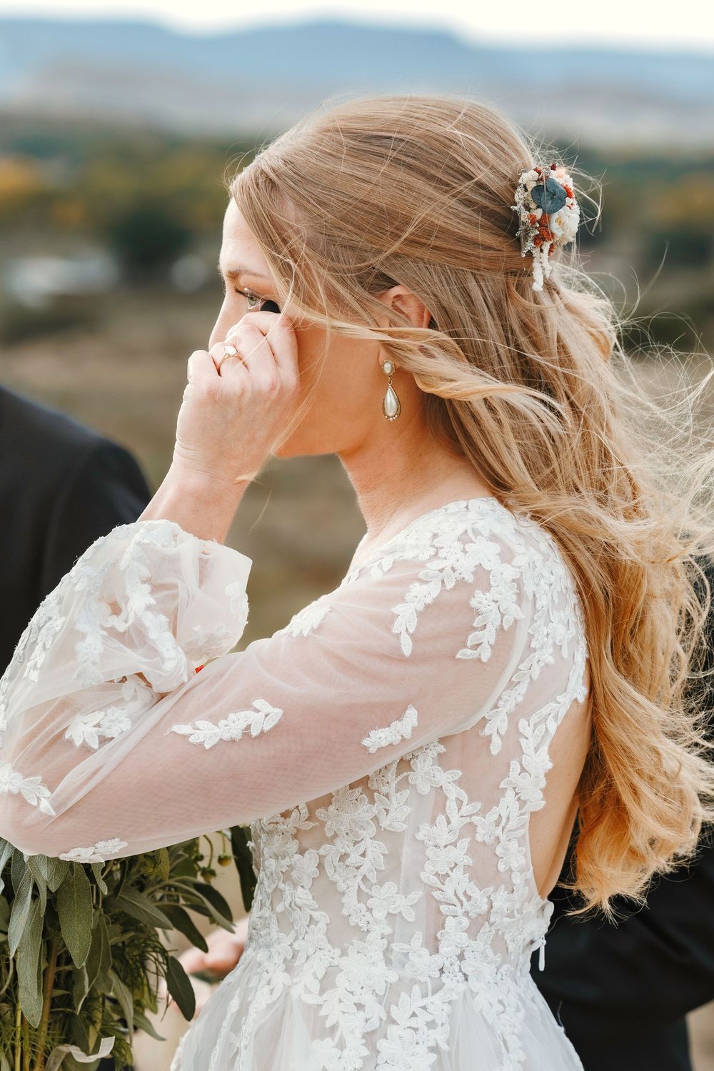 Emotional bride in lace wedding gown covering mouth with hand, floral hair clip and earrings visible, mountains behind.