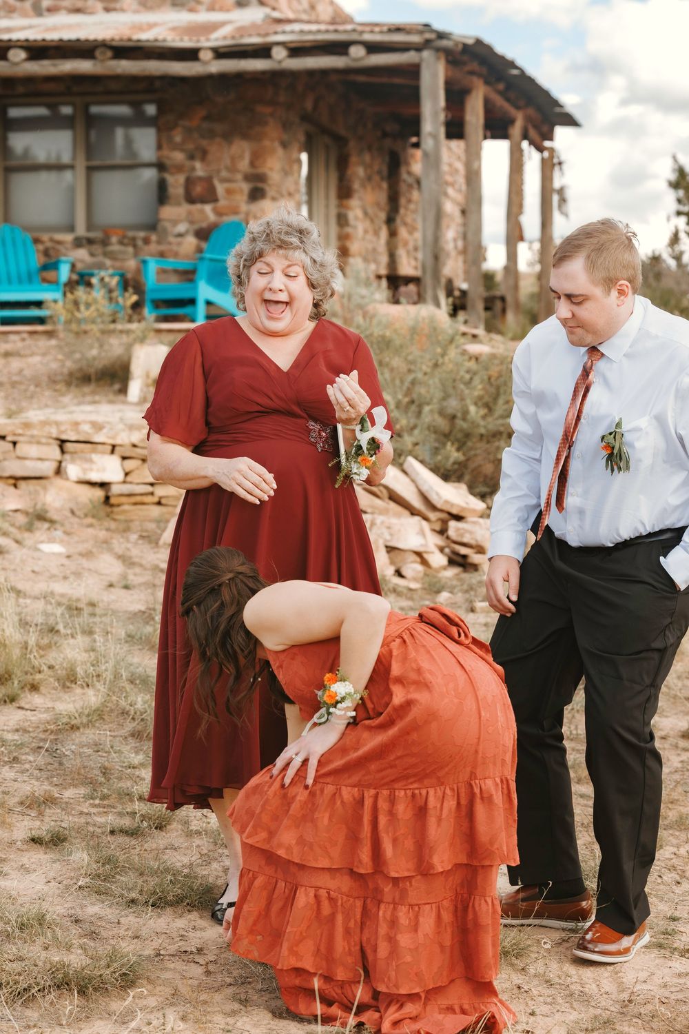 Laughing woman in red dress with corsage at rustic outdoor wedding celebration with stone building backdrop.