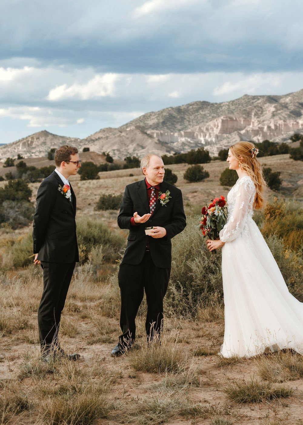 Outdoor wedding ceremony with groom, officiant, and bride holding red bouquet against dramatic mountain landscape.