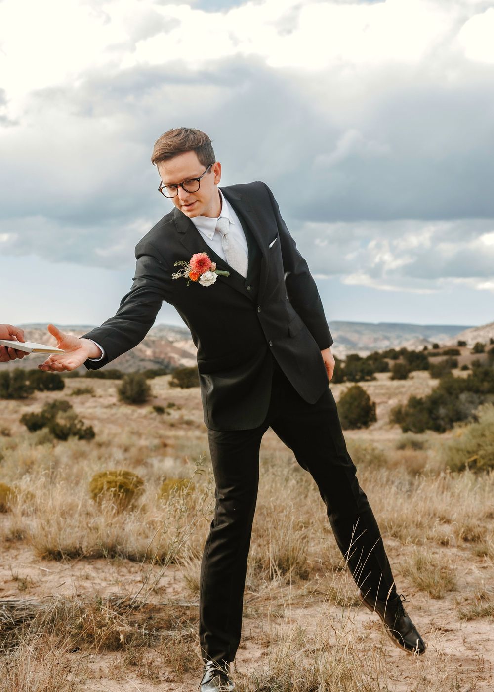 Dapper groom in black suit with floral boutonniere reaching out in arid desert landscape under cloudy sky.
