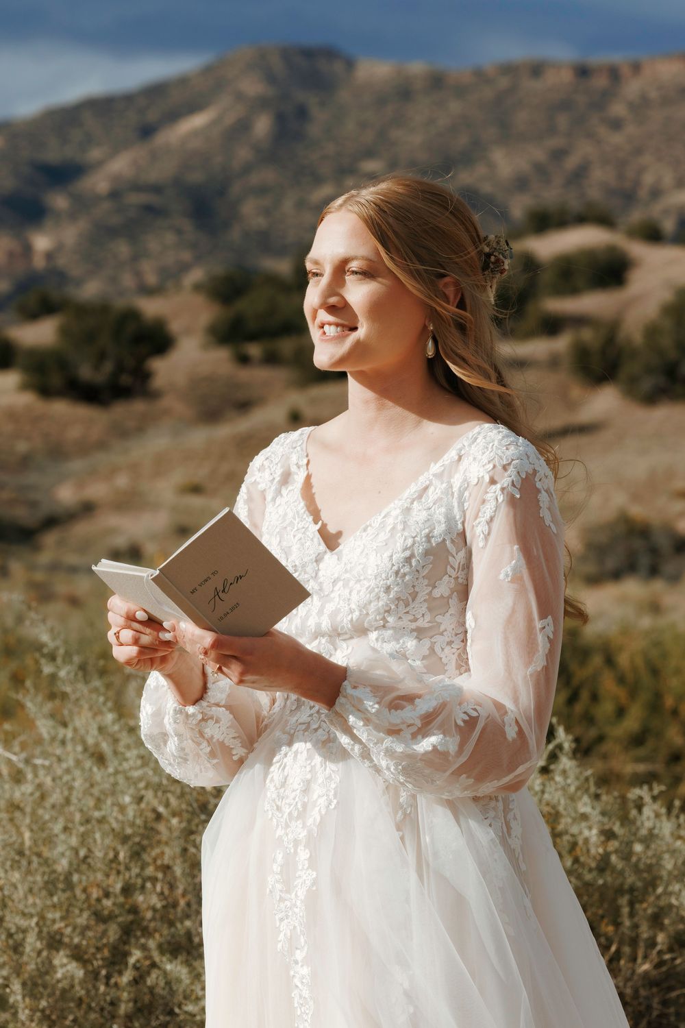 Smiling bride in lace wedding gown reading vows from book with scenic mountain backdrop in sunlight.