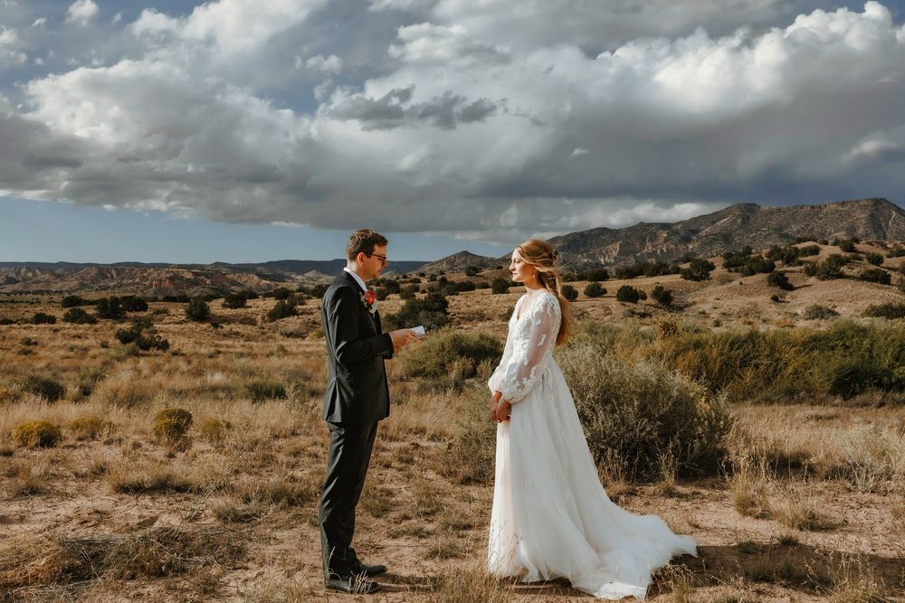 Bride and groom exchanging vows in open desert field with rolling hills and dramatic cloudy sky.