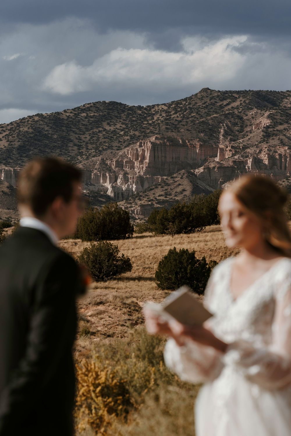 Bride in lace gown reading wedding vows with groom in foreground and dramatic rocky mountain scenery.
