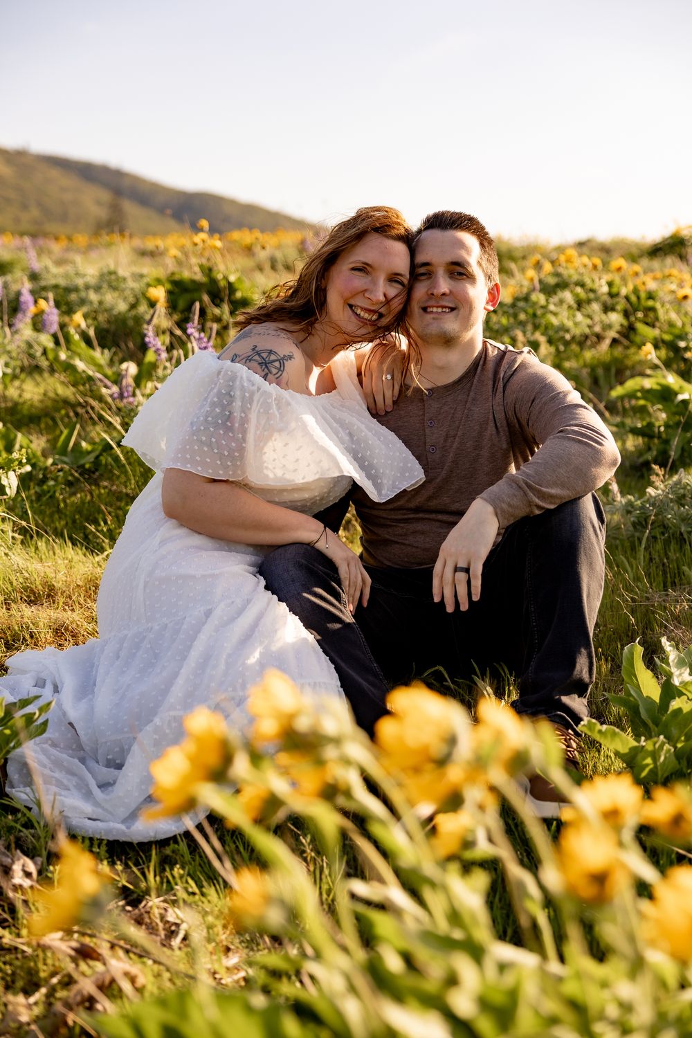 A couple sits together in a meadow of yellow wildflowers during golden hour.