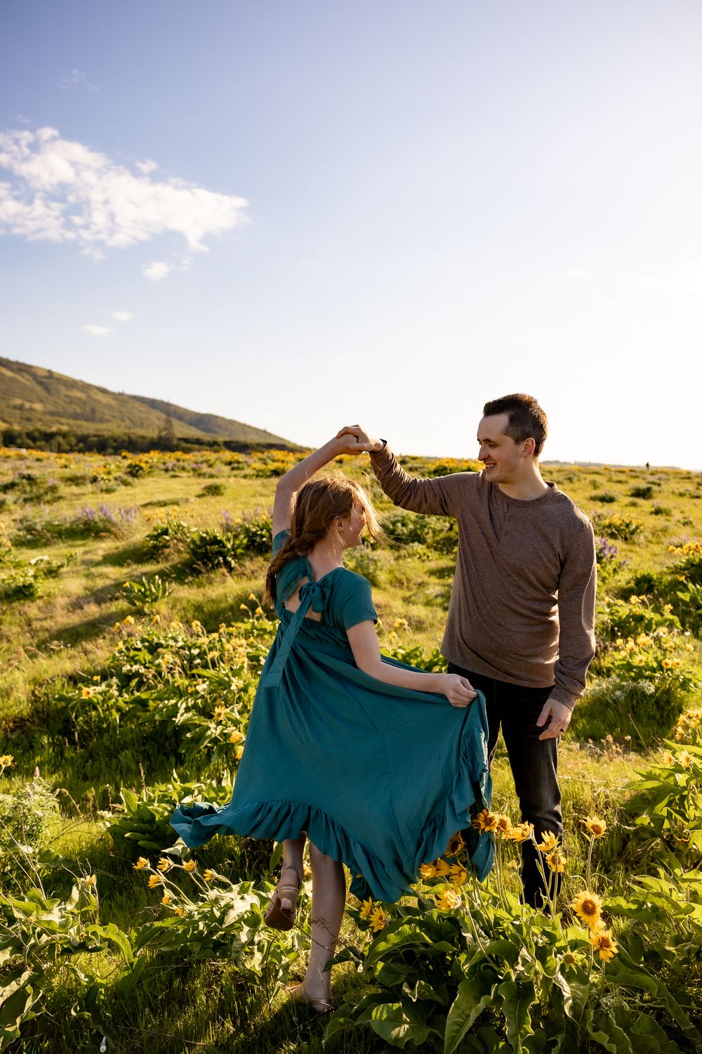 A couple in casual attire share a romantic moment in a sunlit wildflower meadow with mountains in the background.
