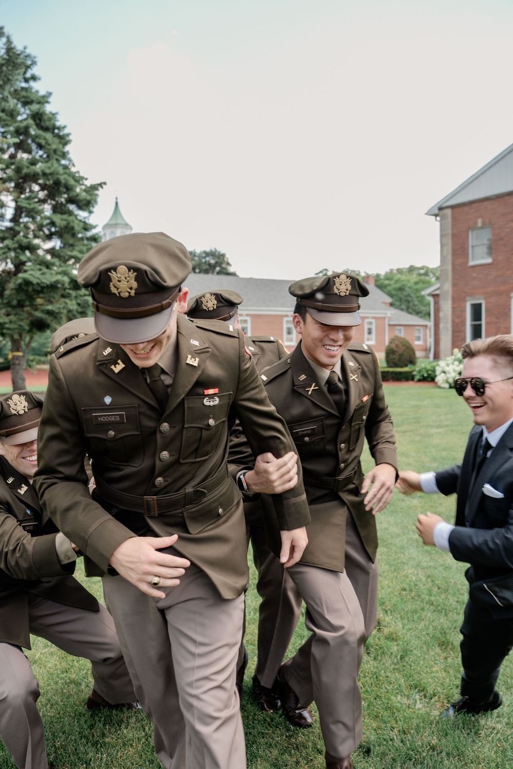 A group in brown military uniforms share a lighthearted moment while bending over on a grassy lawn.