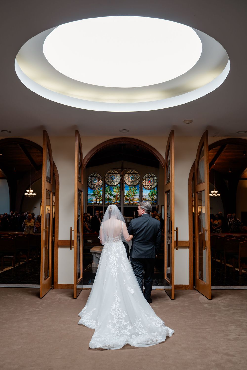 A wedding couple stands beneath circular skylights in an elegant venue with wooden beams and arched windows.