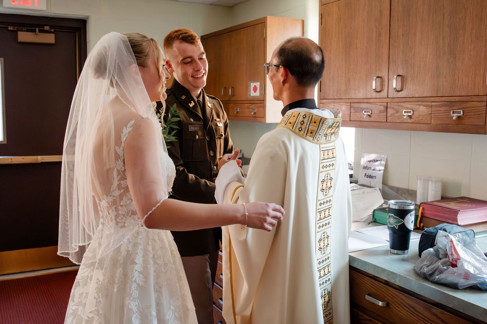 A bride in a white wedding dress gets ready in a rustic wooden kitchen while holding a drink.