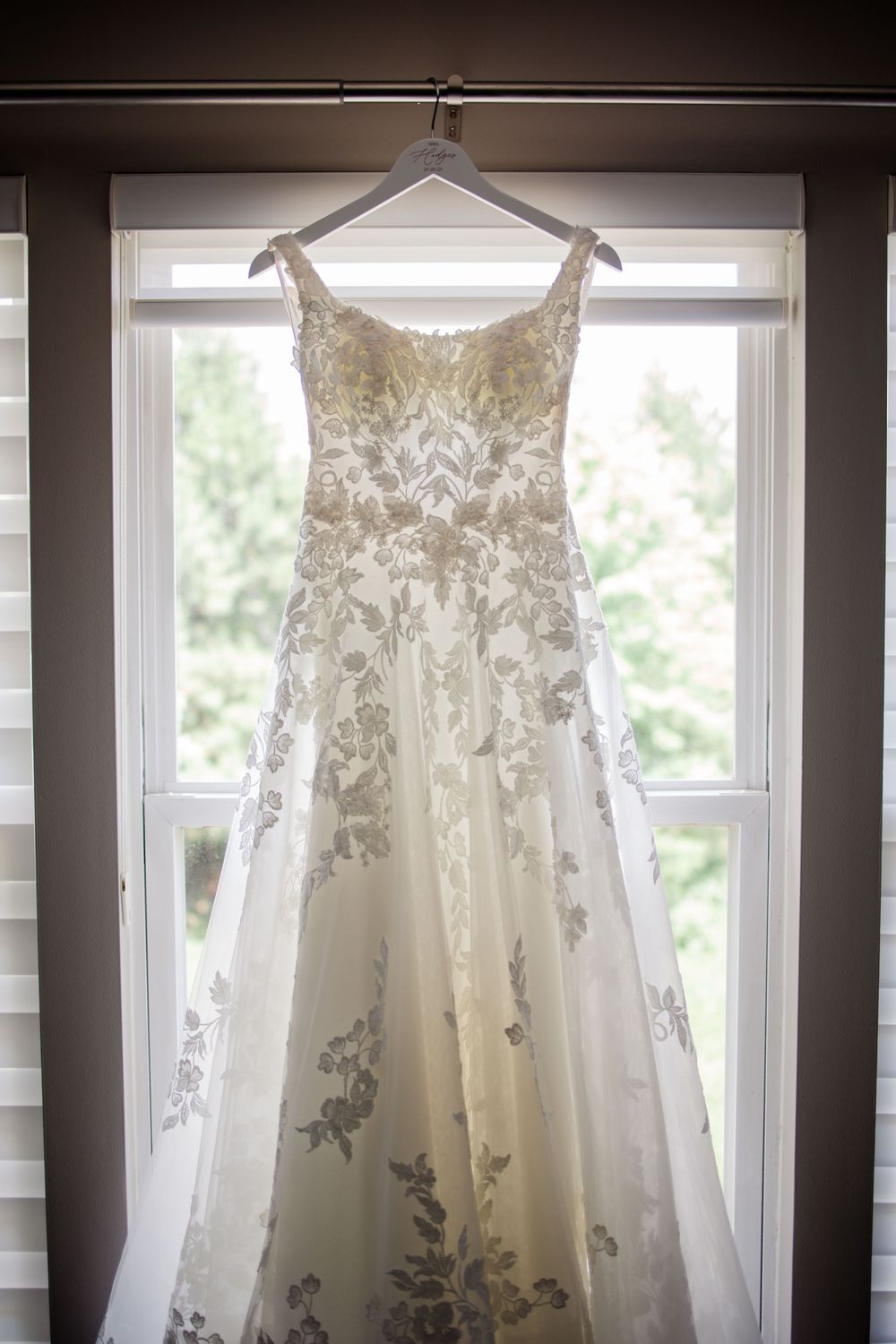 Elegant white lace wedding dress with intricate floral details hanging in front of sunlit windows.
