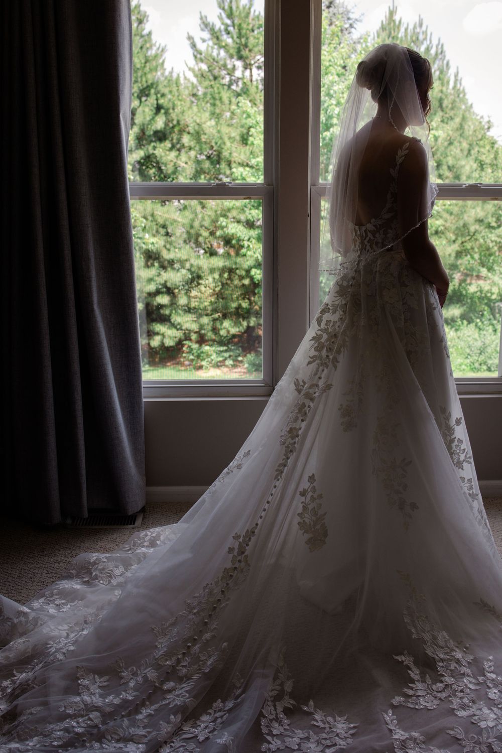 A silhouetted bride in an elegant lace wedding gown with a flowing train stands by a window overlooking pine trees.