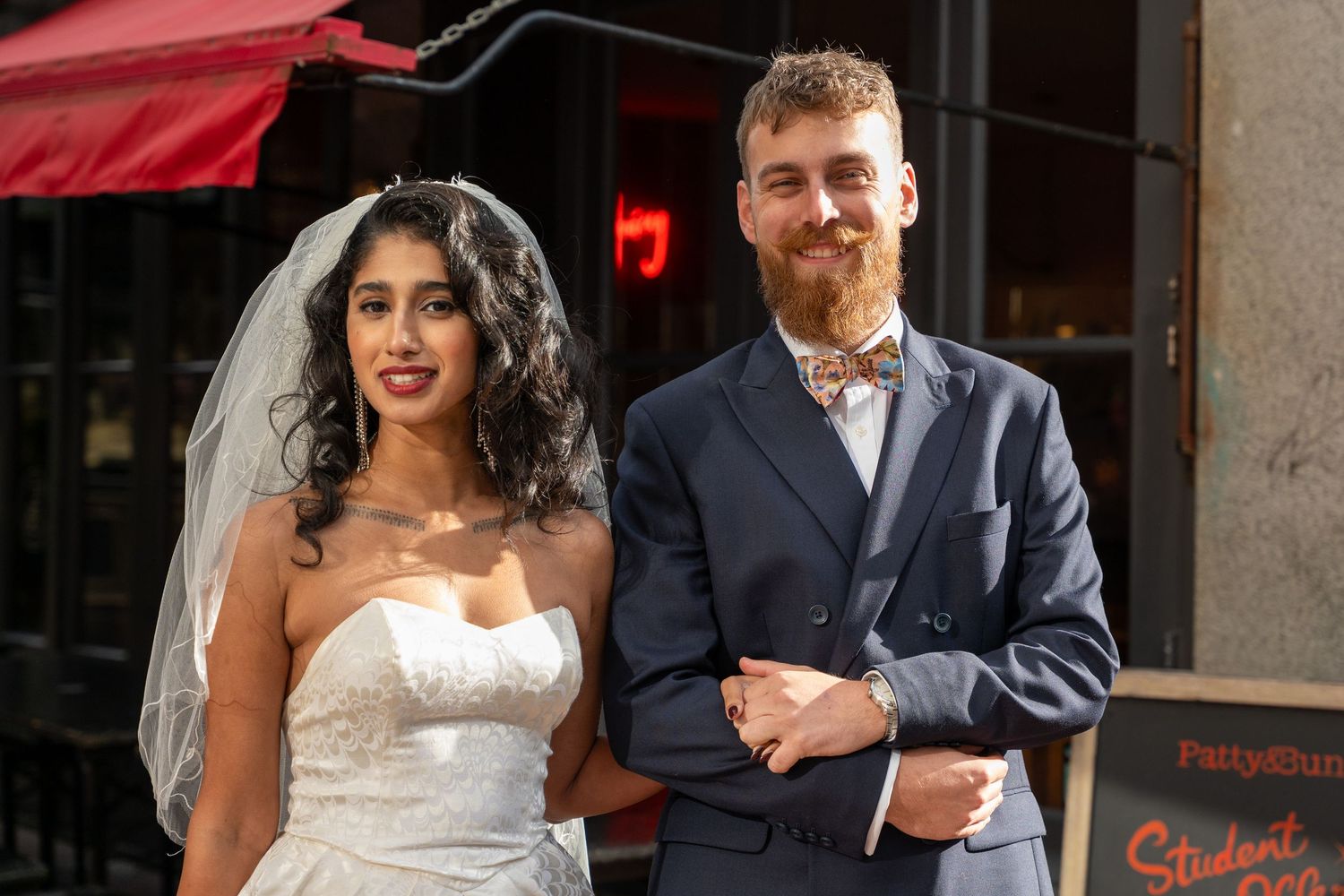 A couple in wedding attire poses together outside a restaurant with a red awning.