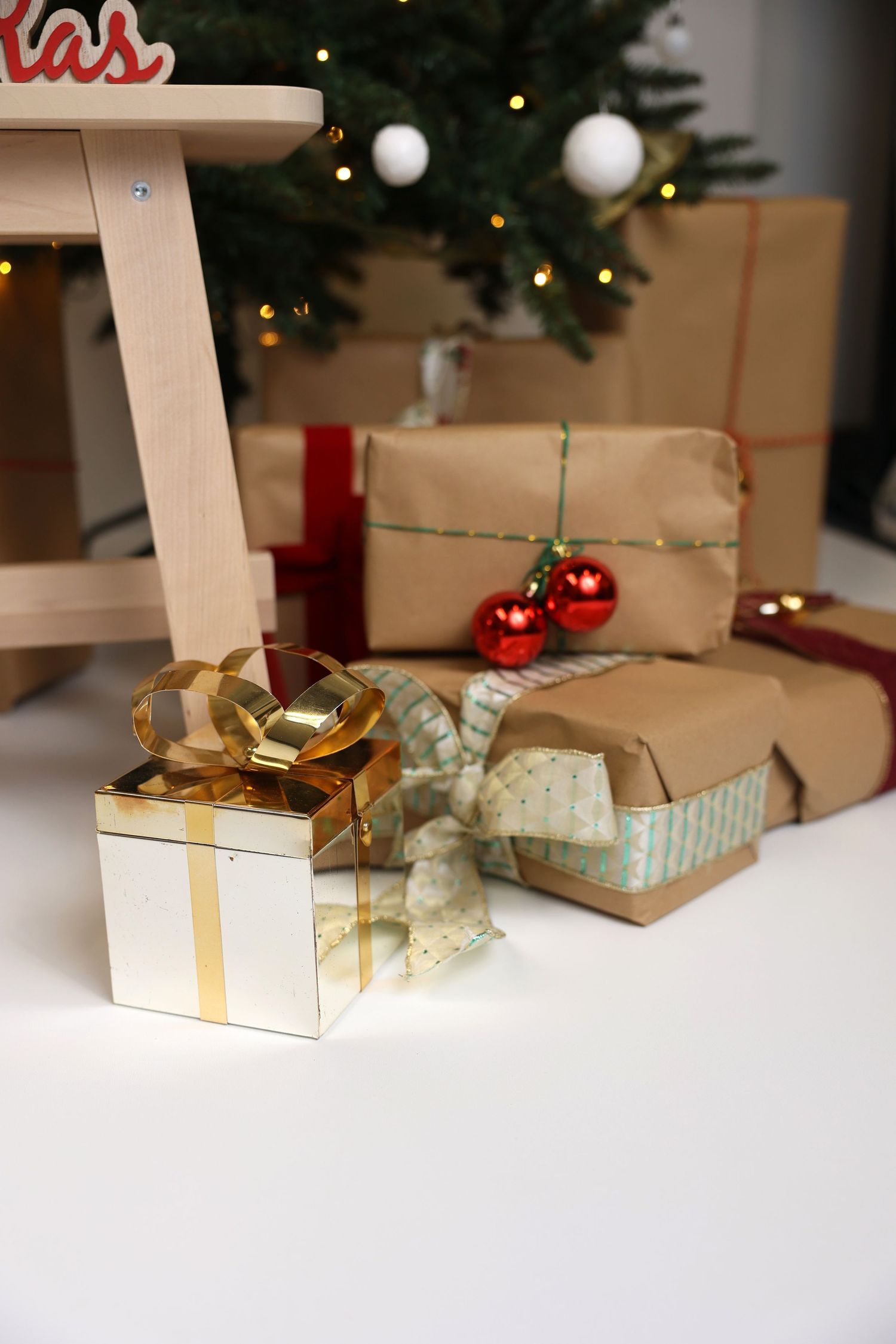 Wrapped Christmas presents with gold and silver bows sit beneath a decorated tree with twinkling lights in the background.