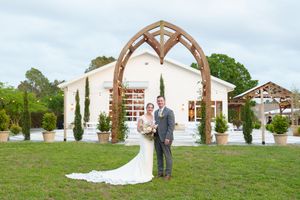 Wedding couple standing under rustic wooden arch with white chapel and cypress trees in background.