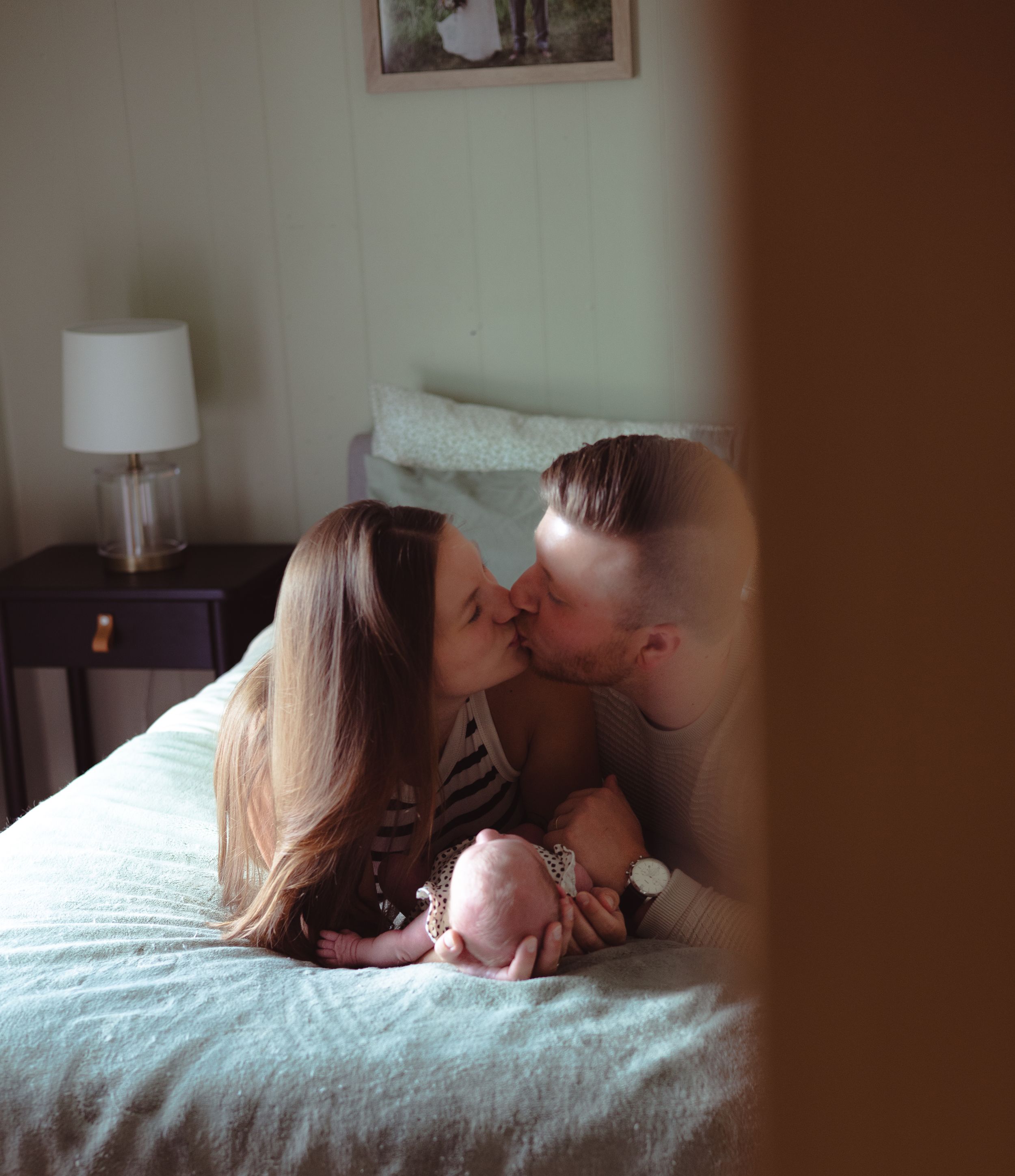 A couple shares an intimate moment while lying together on a bed with white linens in a dimly lit room.