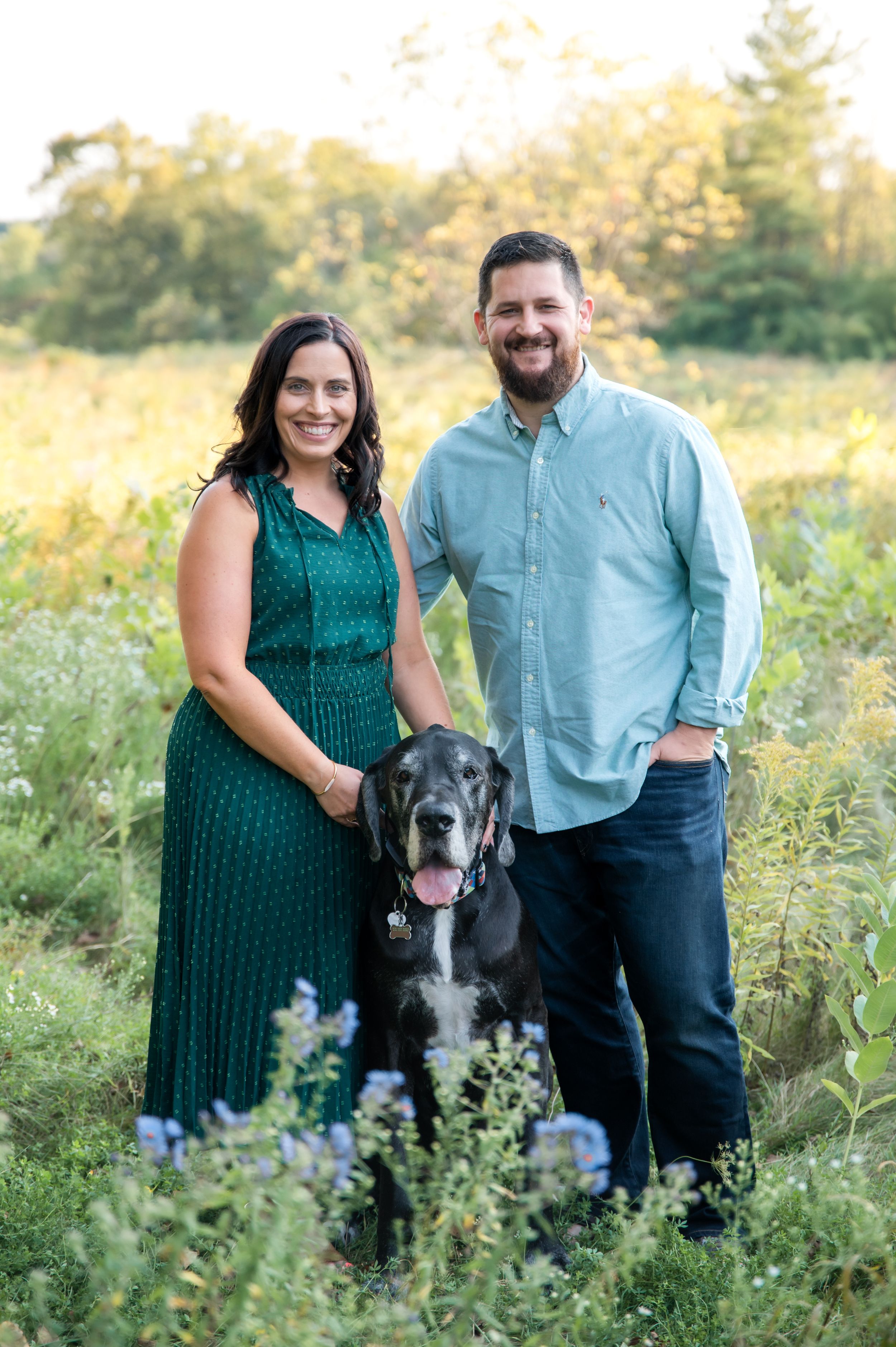 A couple poses with their black dog in a wildflower meadow during golden hour sunset.