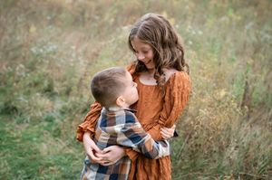 Two children in autumn clothing share a tender moment together in a sunlit meadow with tall grass.