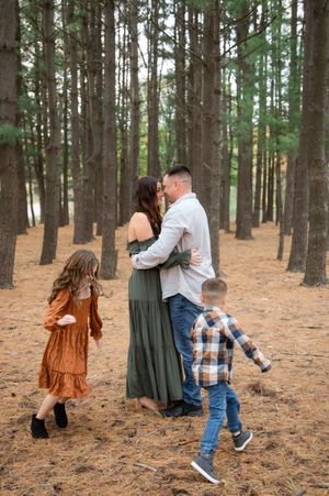 Family plays and dances together among tall pine trees on a forest floor covered in pine needles.