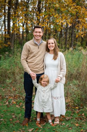 A couple and child in coordinating beige and white outfits pose together in an autumn forest setting.