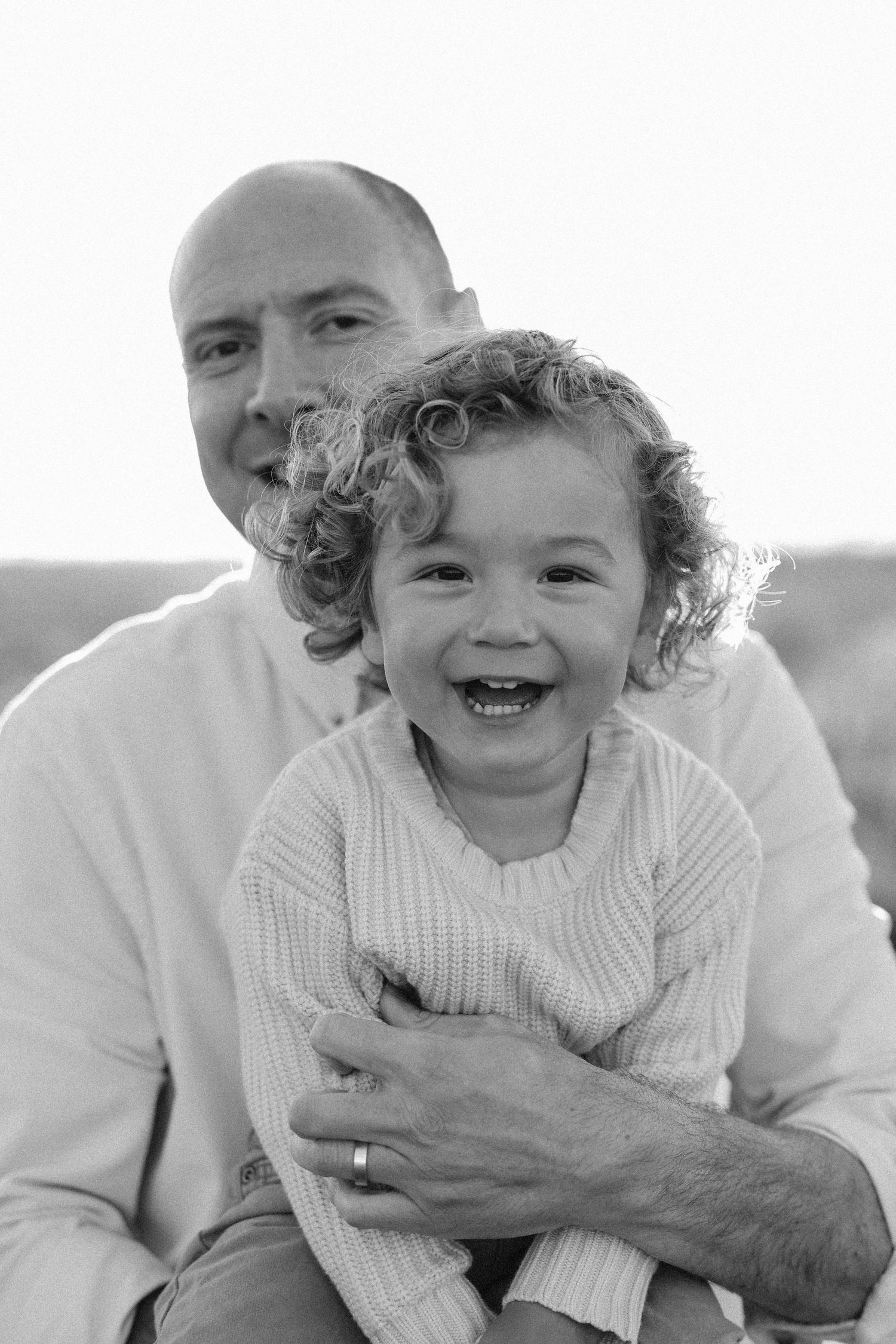 Black and white photo  showing a laughing baby in a white sweater being held and played with dad