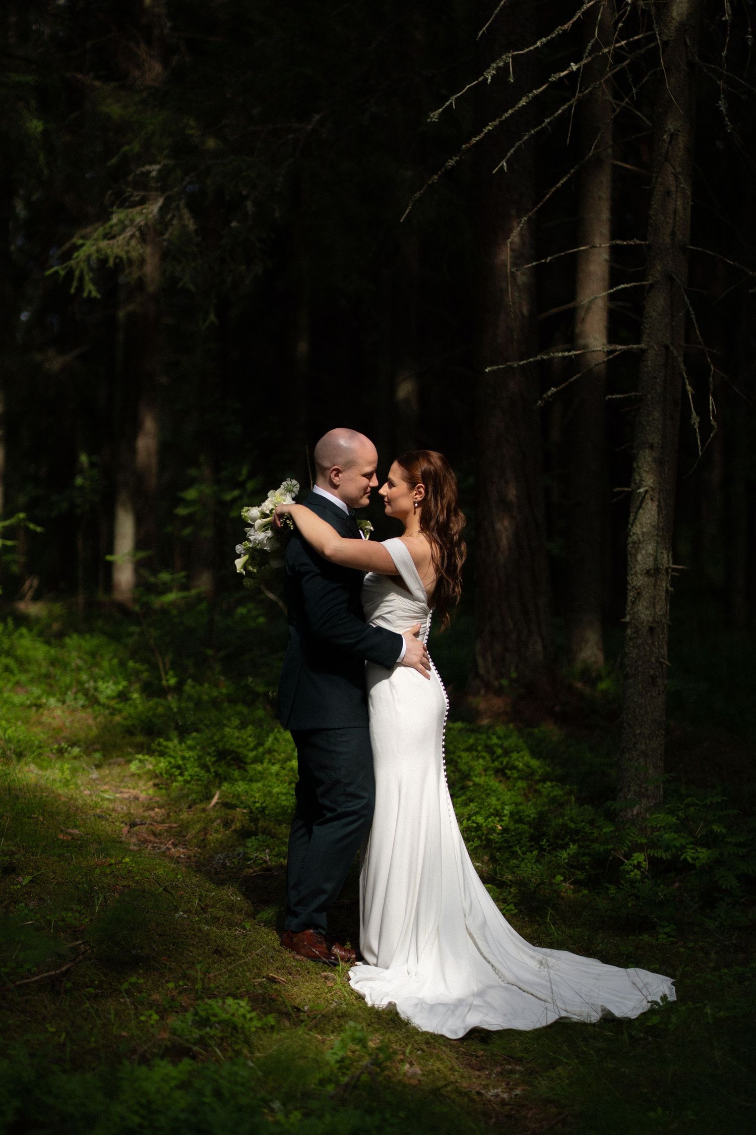 Romantic wedding couple embraces in forest setting with white bouquet and elegant formal attire.
