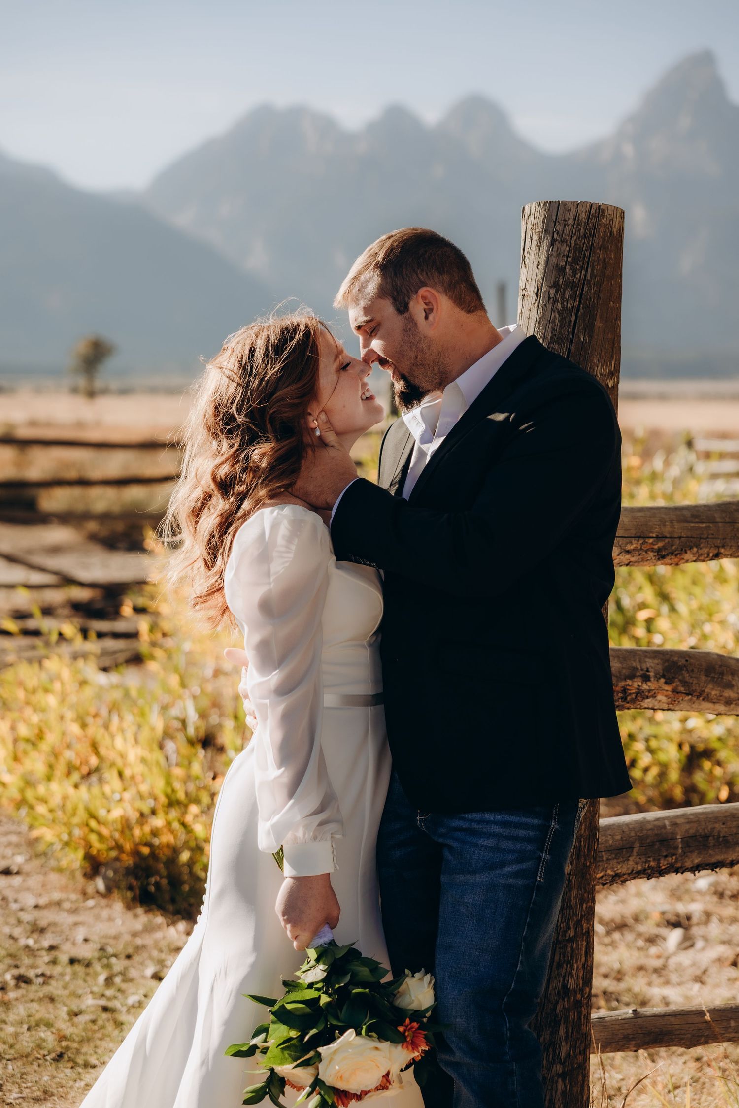 A romantic couple in white embraces tenderly during an outdoor photoshoot at sunset.