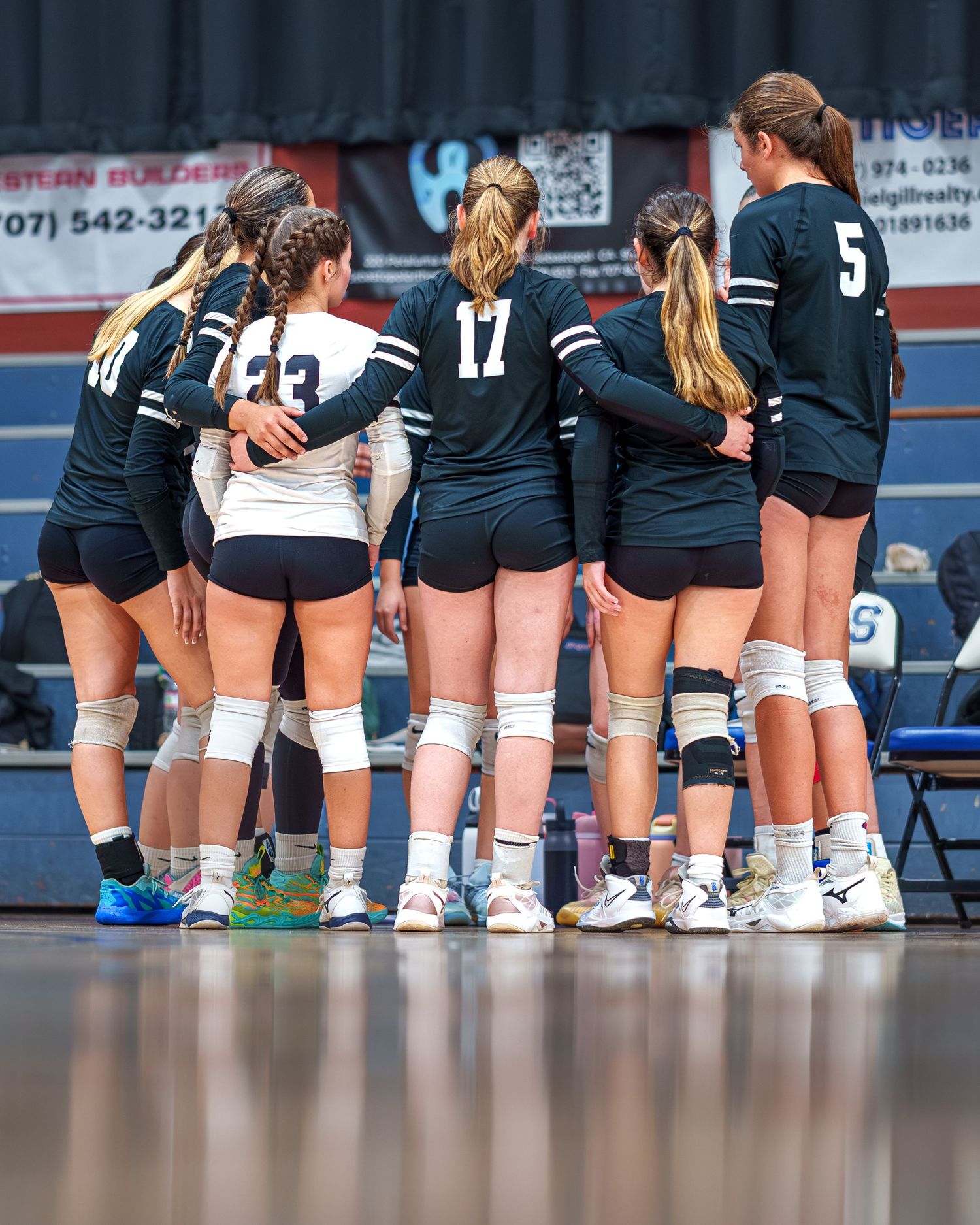 Volleyball team huddles together on court wearing navy blue uniforms with white accents during a match.