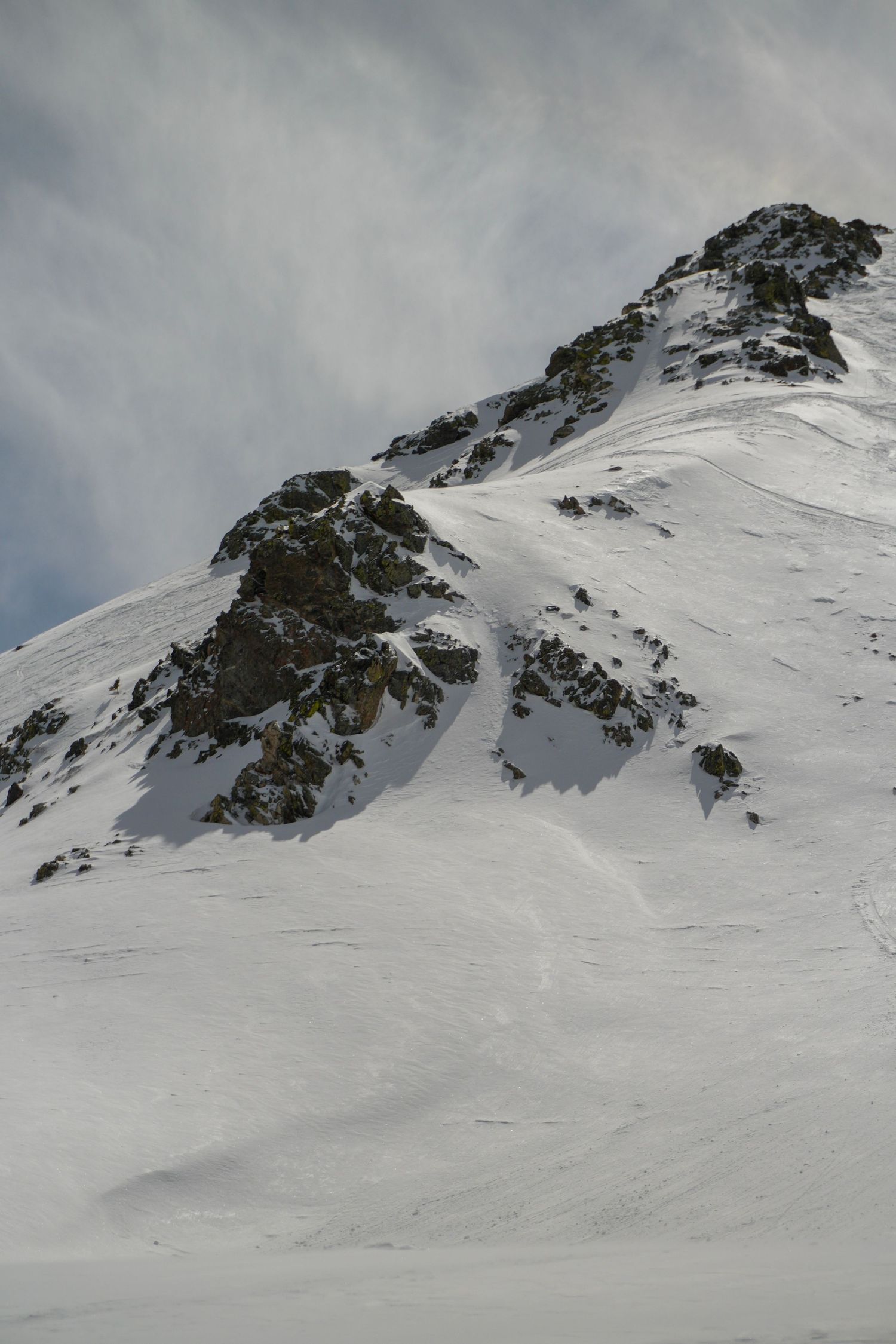 Snow-covered mountain peak with rocky outcrops under a cloudy grey sky.