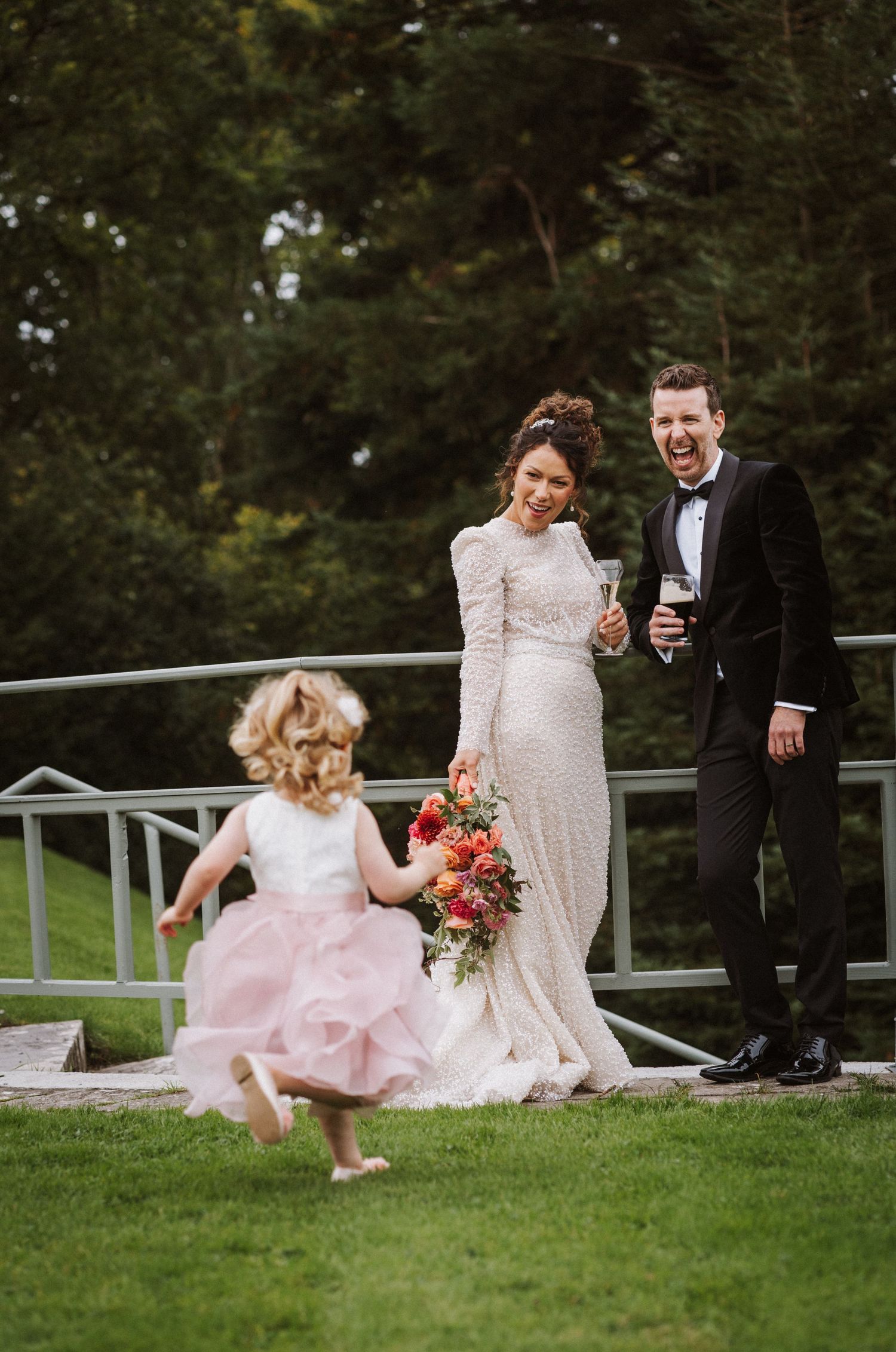 A joyful wedding scene on a lawn with a dancing flower girl in pink while a couple shares a laugh by the railing.