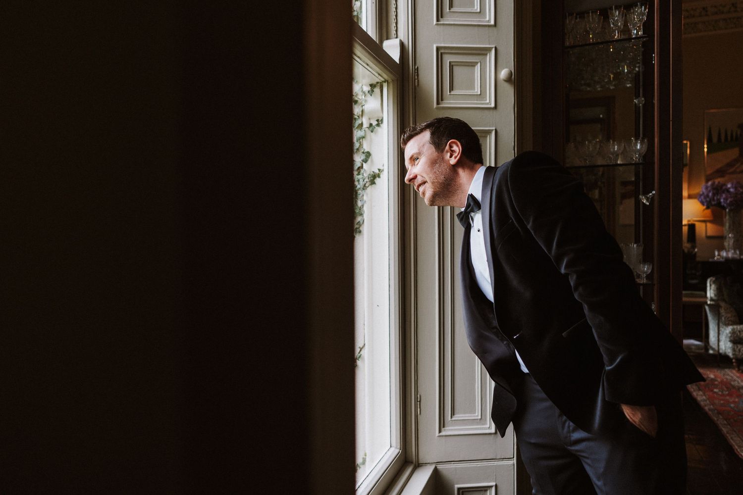 A groom in a black tuxedo leans against an elegant white window frame in a classic wedding portrait.