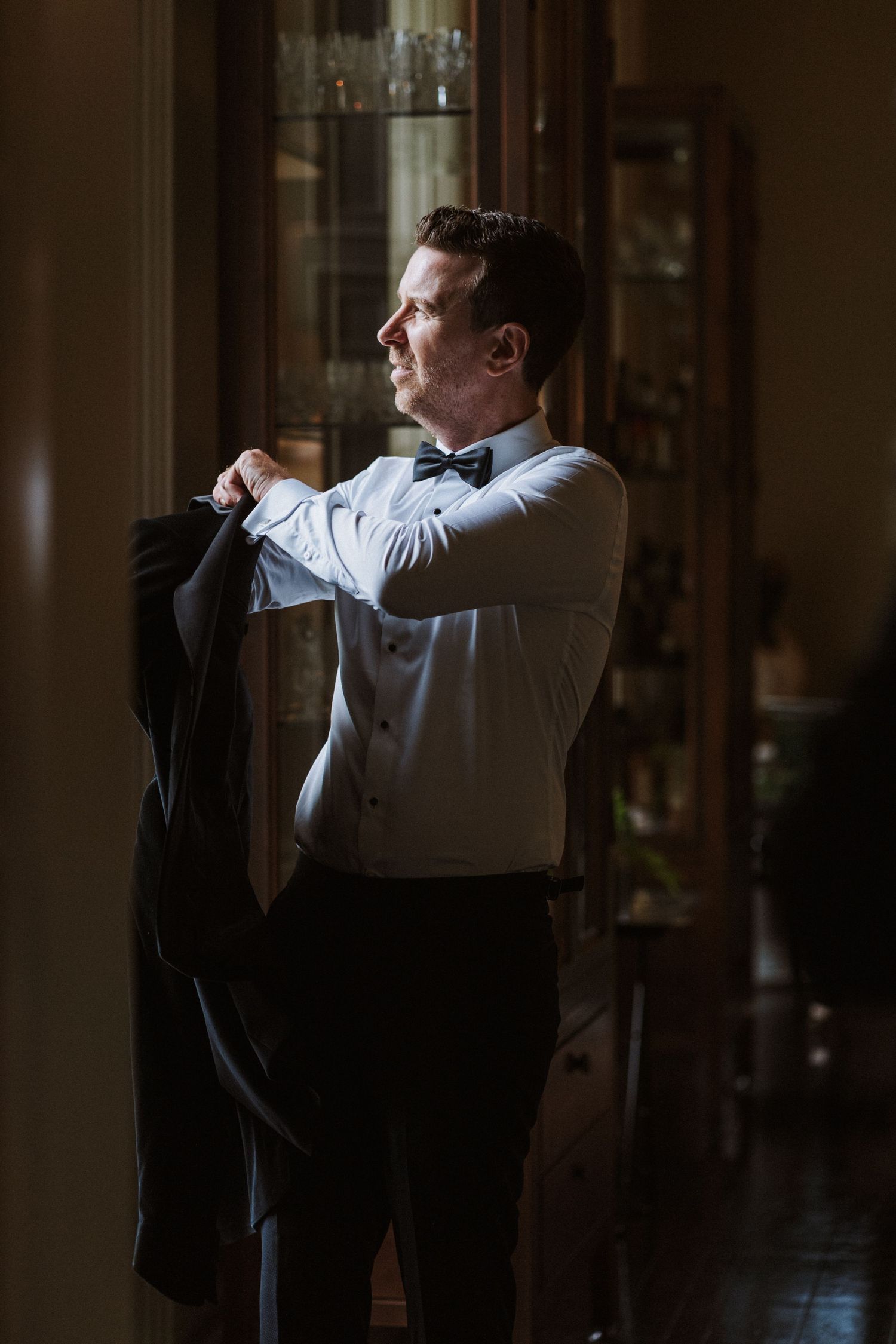 A person in formal attire adjusts their bowtie while getting dressed in a dimly lit room.