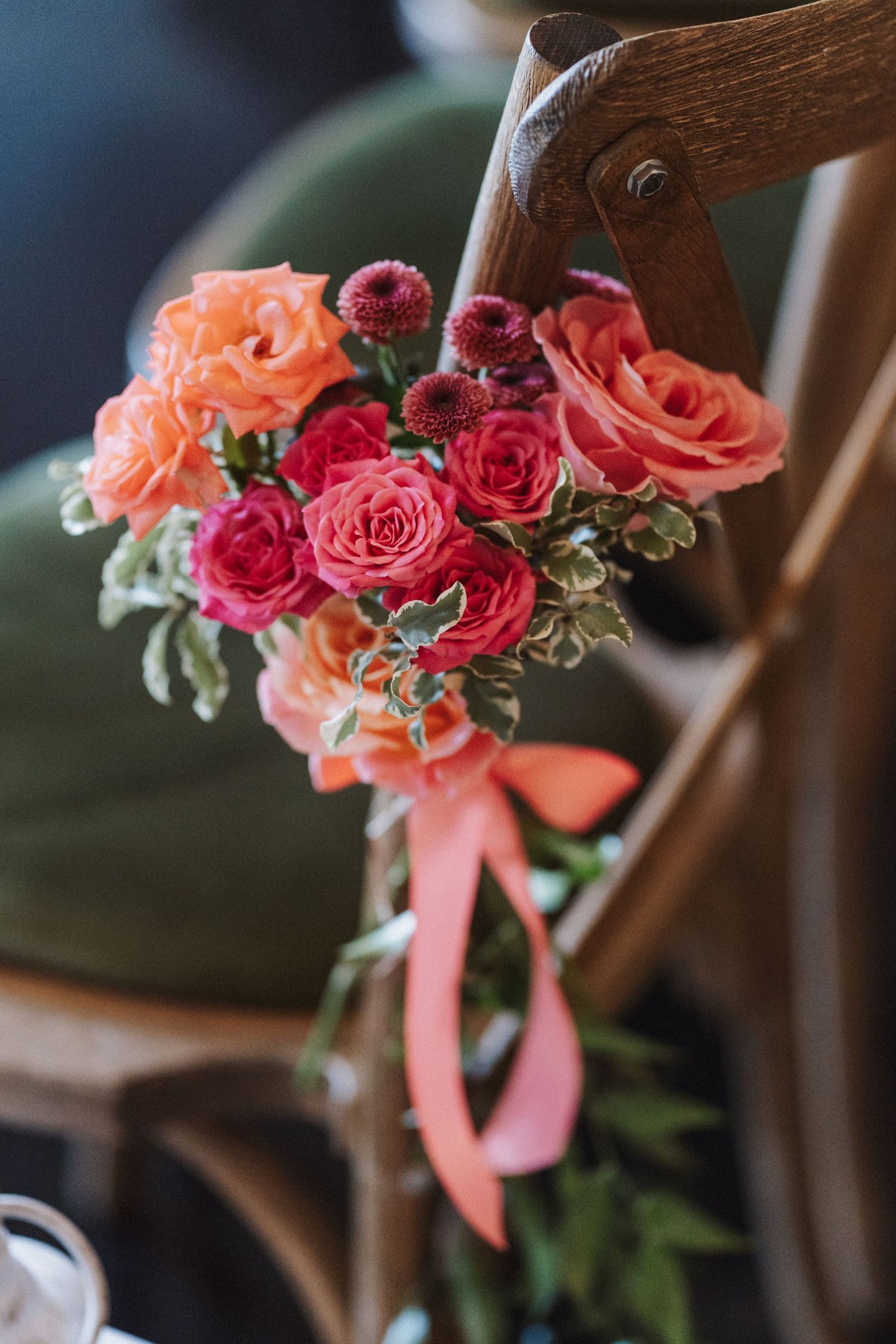 Coral and red roses with greenery arranged on wooden chairs with peach ribbons for wedding ceremony decor.